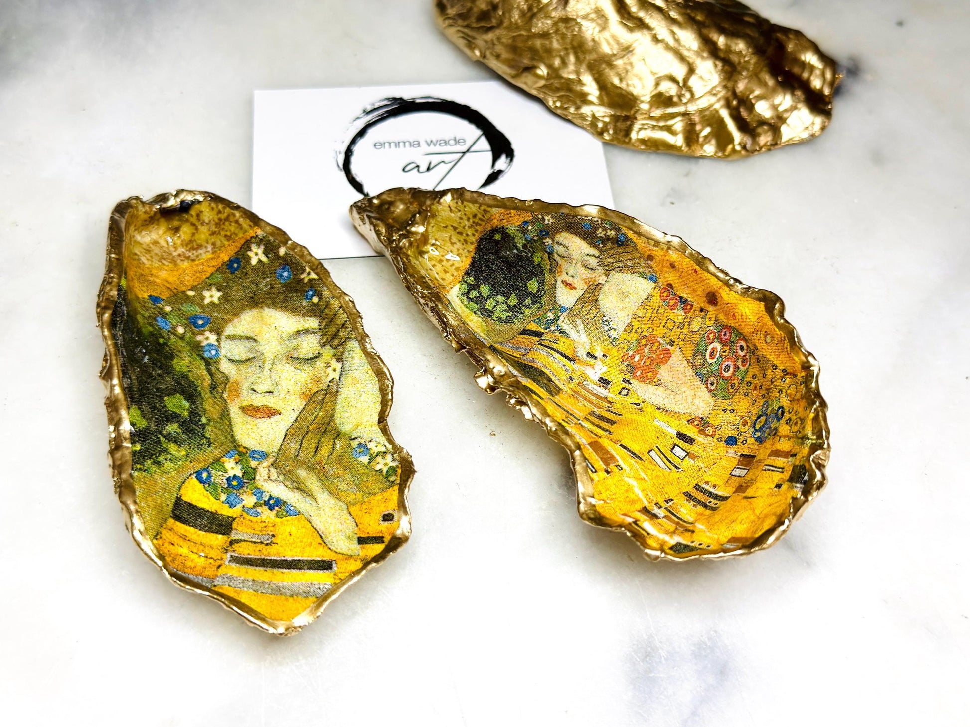 Handcrafted Dorset oyster shell trinket dish inspired by Gustav Klimt’s The Kiss, featuring romantic gold and green artwork detail. Natural oyster shell edged in hand applied liquid gold leaf and sealed with high gloss resin. Luxury art inspired jewellery holder displayed with gold rings on marble background. Unique gift for her or art lover keepsake.