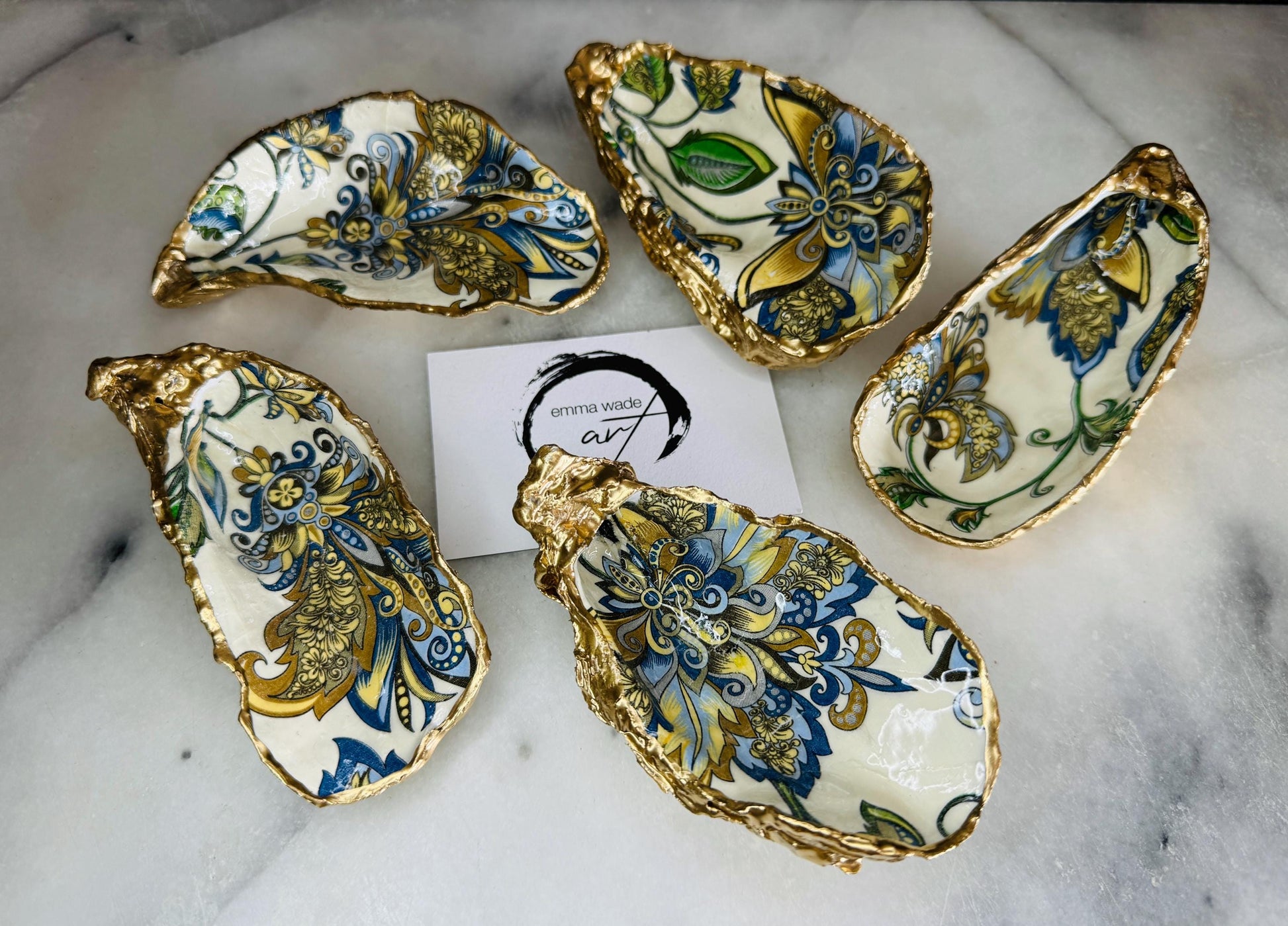 Handmade Dorset oyster shell trinket dishes decorated with ornate blue, green and gold baroque floral patterns inspired by antique European porcelain and vintage decorative art. Natural oyster shells are hand gilded with liquid gold leaf edging and sealed with glossy crystal clear resin. Luxury coastal jewelry holders and vanity dishes photographed on marble background with elegant decorative styling.