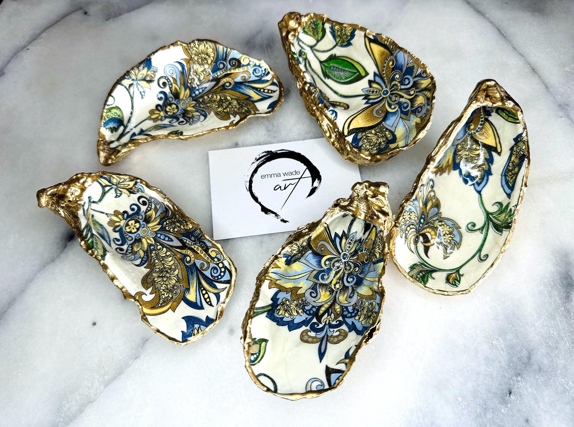Handmade Dorset oyster shell trinket dishes decorated with ornate blue, green and gold baroque floral patterns inspired by antique European porcelain and vintage decorative art. Natural oyster shells are hand gilded with liquid gold leaf edging and sealed with glossy crystal clear resin. Luxury coastal jewelry holders and vanity dishes photographed on marble background with elegant decorative styling.