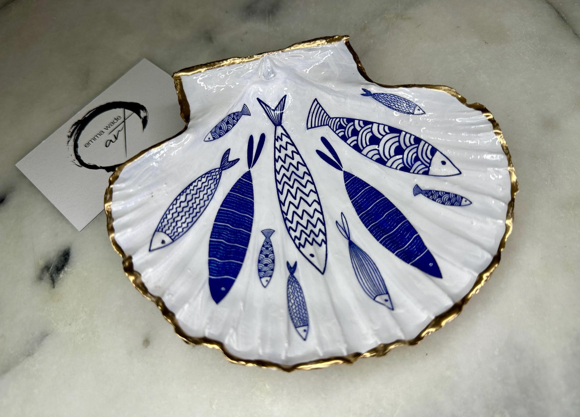 Handmade Dorset scallop shell trinket dishes featuring elegant blue fish illustrations with hand-painted gold leaf edging and glossy crystal clear resin. Coastal nautical jewellery dishes inspired by Scandinavian ocean decor, styled on a neutral background with detailed blue fish artwork and luxurious gilded edges.