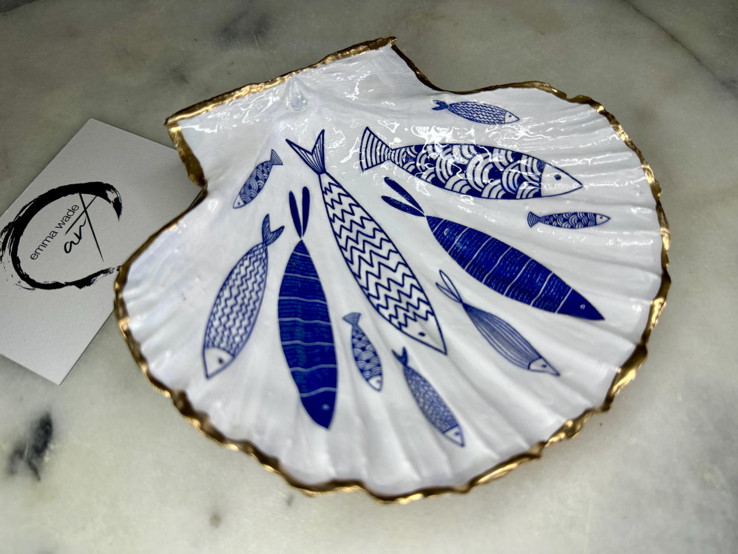 Handmade Dorset scallop shell trinket dishes featuring elegant blue fish illustrations with hand-painted gold leaf edging and glossy crystal clear resin. Coastal nautical jewellery dishes inspired by Scandinavian ocean decor, styled on a neutral background with detailed blue fish artwork and luxurious gilded edges.