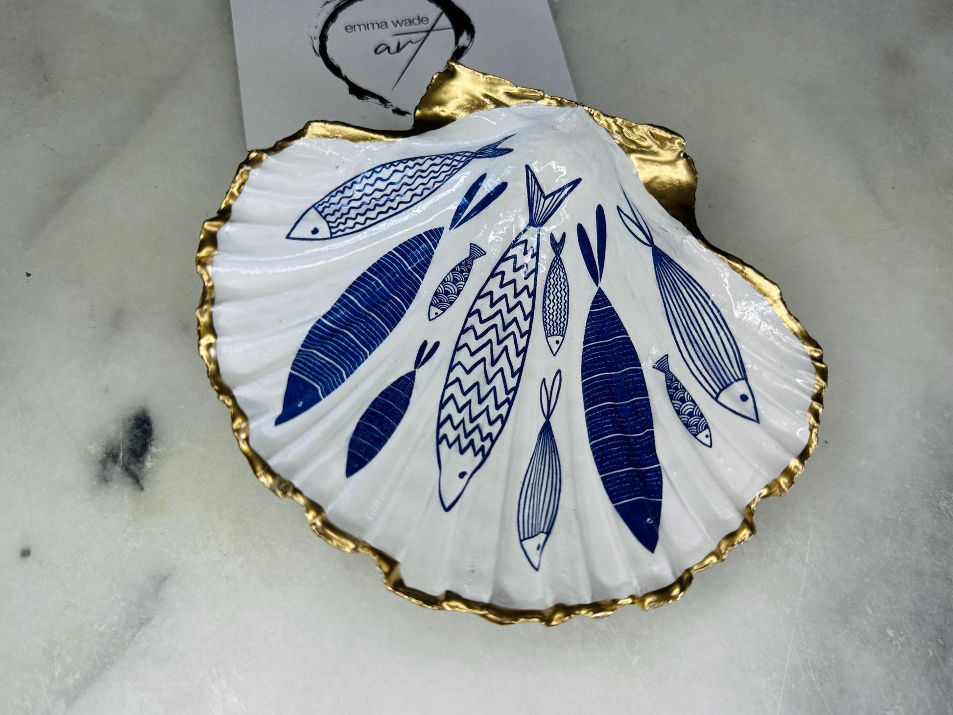 Handmade Dorset scallop shell trinket dishes featuring elegant blue fish illustrations with hand-painted gold leaf edging and glossy crystal clear resin. Coastal nautical jewellery dishes inspired by Scandinavian ocean decor, styled on a neutral background with detailed blue fish artwork and luxurious gilded edges.