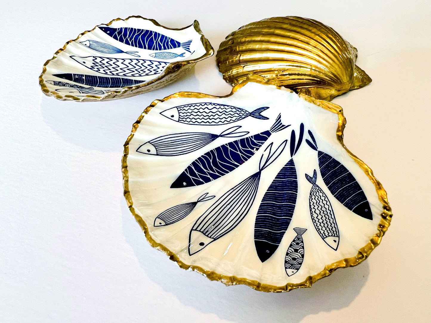 Handmade Dorset scallop shell trinket dishes featuring elegant blue fish illustrations with hand-painted gold leaf edging and glossy crystal clear resin. Coastal nautical jewellery dishes inspired by Scandinavian ocean decor, styled on a neutral background with detailed blue fish artwork and luxurious gilded edges.