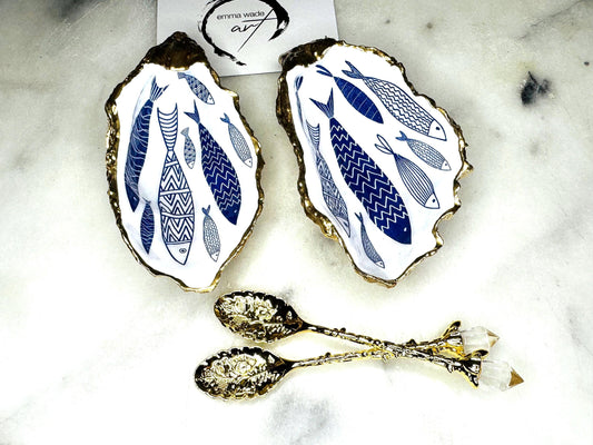 Pair of handmade Dorset oyster shell salt and pepper pinch pots decorated with elegant blue and white fish artwork, finished with hand applied liquid gold leaf edging and sealed in glossy food-safe resin. Coastal shell cruet dishes styled with optional gold crystal spoons on a marble background. Luxury nautical kitchen decor inspired by the sea, perfect for beach house dining, dinner parties, weddings, anniversaries, and coastal home gifts.
