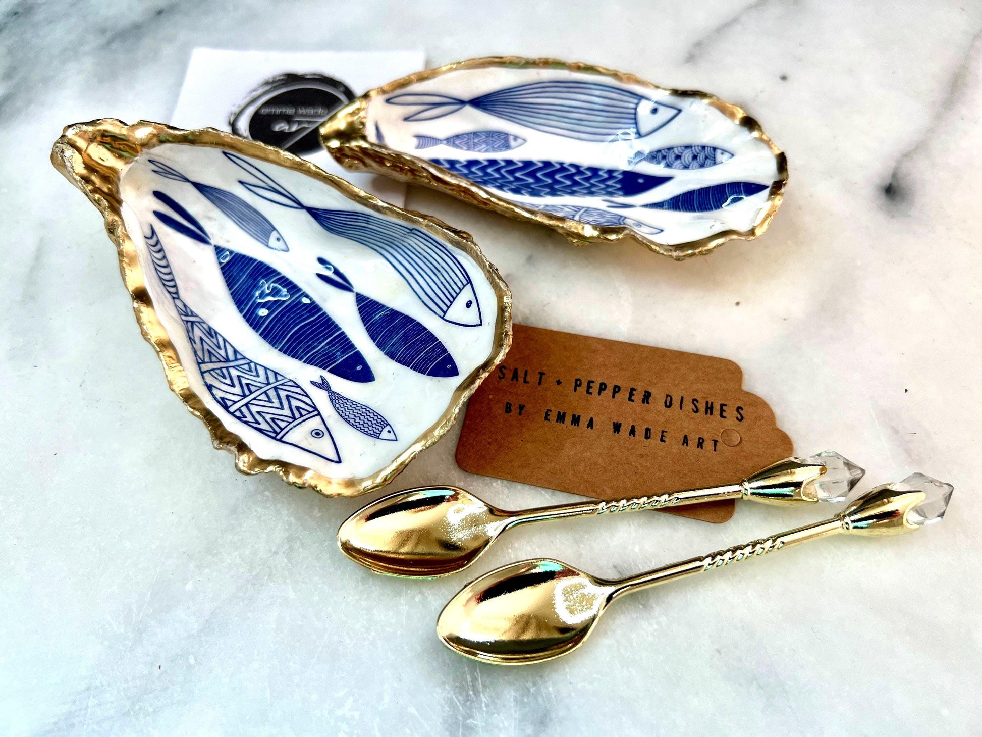 Pair of handmade Dorset oyster shell salt and pepper pinch pots decorated with elegant blue and white fish artwork, finished with hand applied liquid gold leaf edging and sealed in glossy food-safe resin. Coastal shell cruet dishes styled with optional gold crystal spoons on a marble background. Luxury nautical kitchen decor inspired by the sea, perfect for beach house dining, dinner parties, weddings, anniversaries, and coastal home gifts.