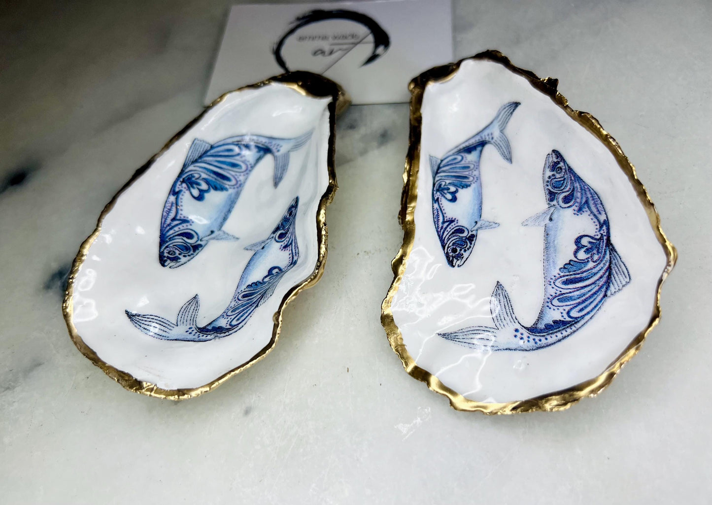 Handmade Dorset oyster shell trinket dish with blue and white paisley fish design, gold leaf gilded edges and glossy resin finish. Coastal jewellery holder and ring dish, photographed on a light surface showing the natural shell shape and pearly interior. Unique sustainably sourced shell, sold individually as a decorative coastal home décor accent or gift.