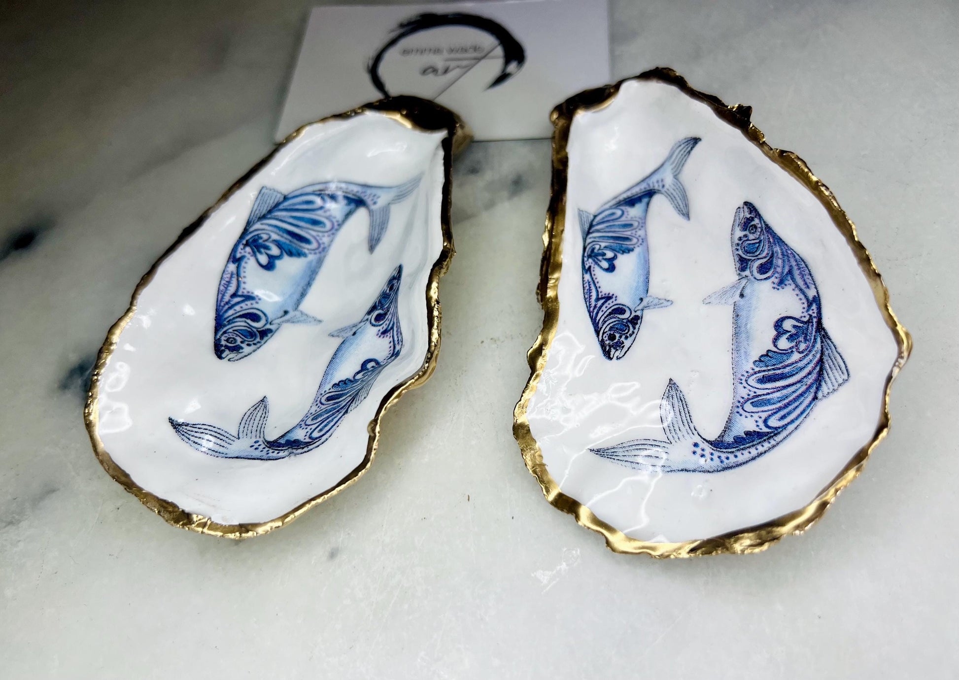 Handmade Dorset oyster shell trinket dish with blue and white paisley fish design, gold leaf gilded edges and glossy resin finish. Coastal jewellery holder and ring dish, photographed on a light surface showing the natural shell shape and pearly interior. Unique sustainably sourced shell, sold individually as a decorative coastal home décor accent or gift.