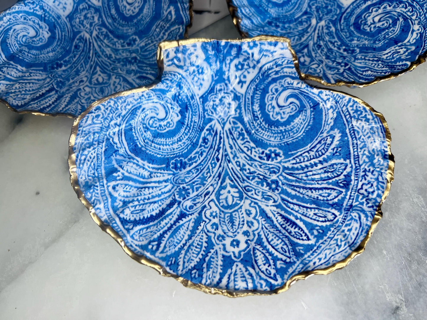 Blue and white paisley scallop shell ring catcher with hand applied gold leaf edging and glossy resin finish. Handmade Dorset shell jewellery tray styled with pearls on a marble background. Coastal vanity decor and luxury beach house accessory gift.