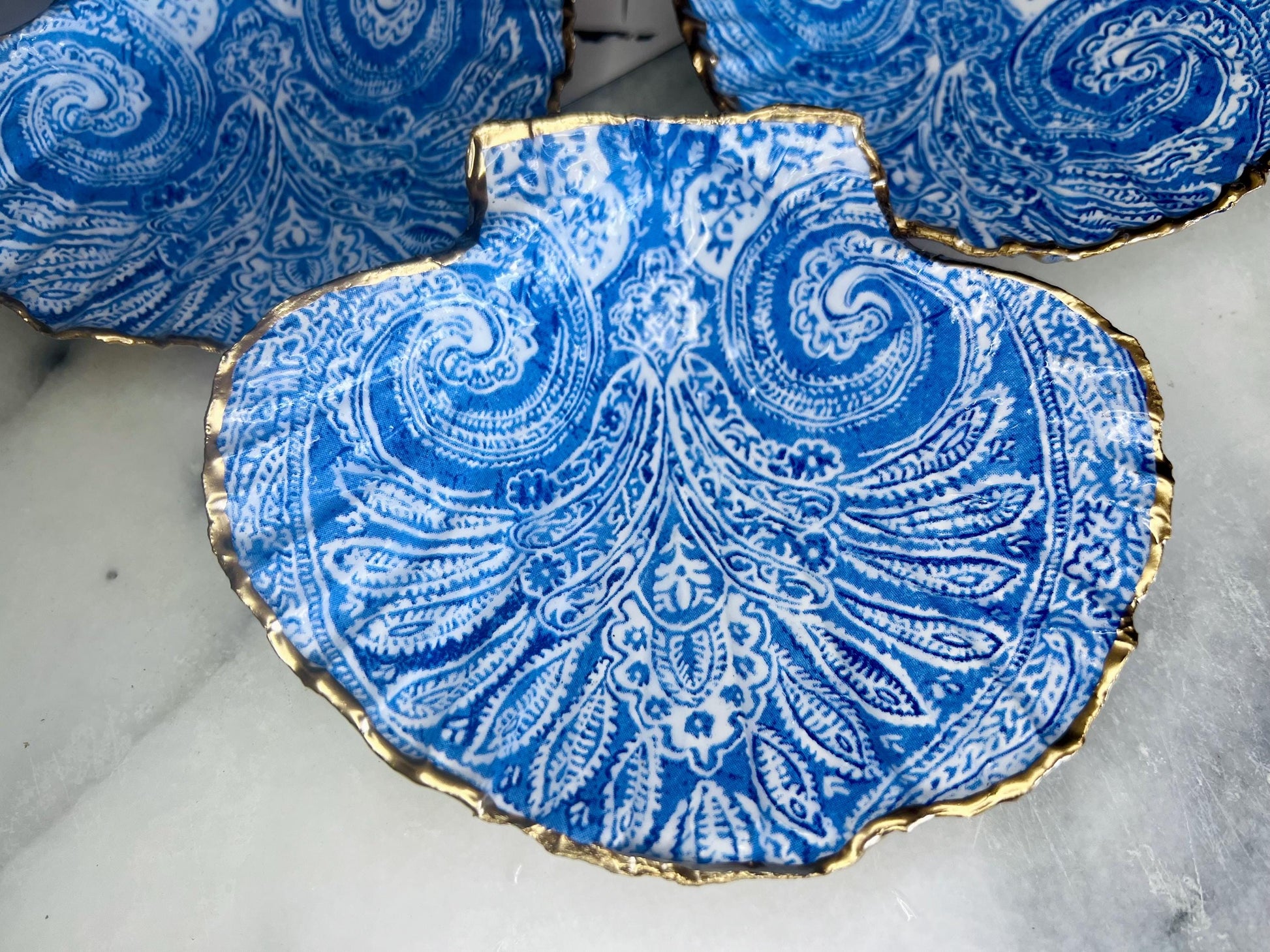 Blue and white paisley scallop shell ring catcher with hand applied gold leaf edging and glossy resin finish. Handmade Dorset shell jewellery tray styled with pearls on a marble background. Coastal vanity decor and luxury beach house accessory gift.