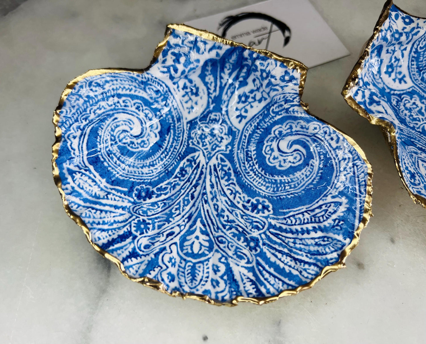 Blue and white paisley scallop shell ring catcher with hand applied gold leaf edging and glossy resin finish. Handmade Dorset shell jewellery tray styled with pearls on a marble background. Coastal vanity decor and luxury beach house accessory gift.