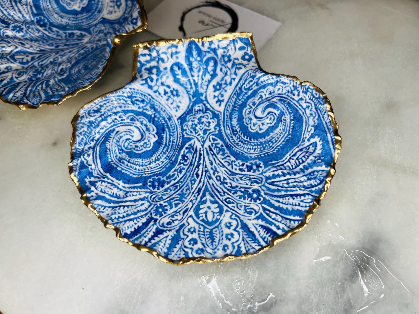 Blue and white paisley scallop shell ring catcher with hand applied gold leaf edging and glossy resin finish. Handmade Dorset shell jewellery tray styled with pearls on a marble background. Coastal vanity decor and luxury beach house accessory gift.