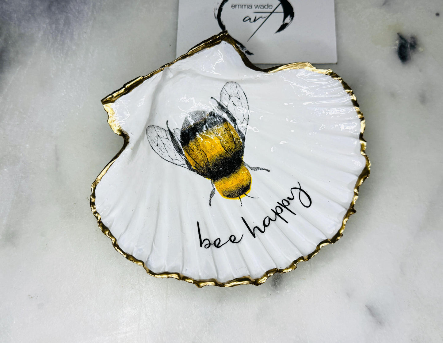 Handmade scallop shell trinket dish featuring a bumblebee illustration with gold leaf edging and glossy resin finish. Coastal jewellery dish in natural white shell with bee artwork, displayed as a ring holder and handmade bee lover gift