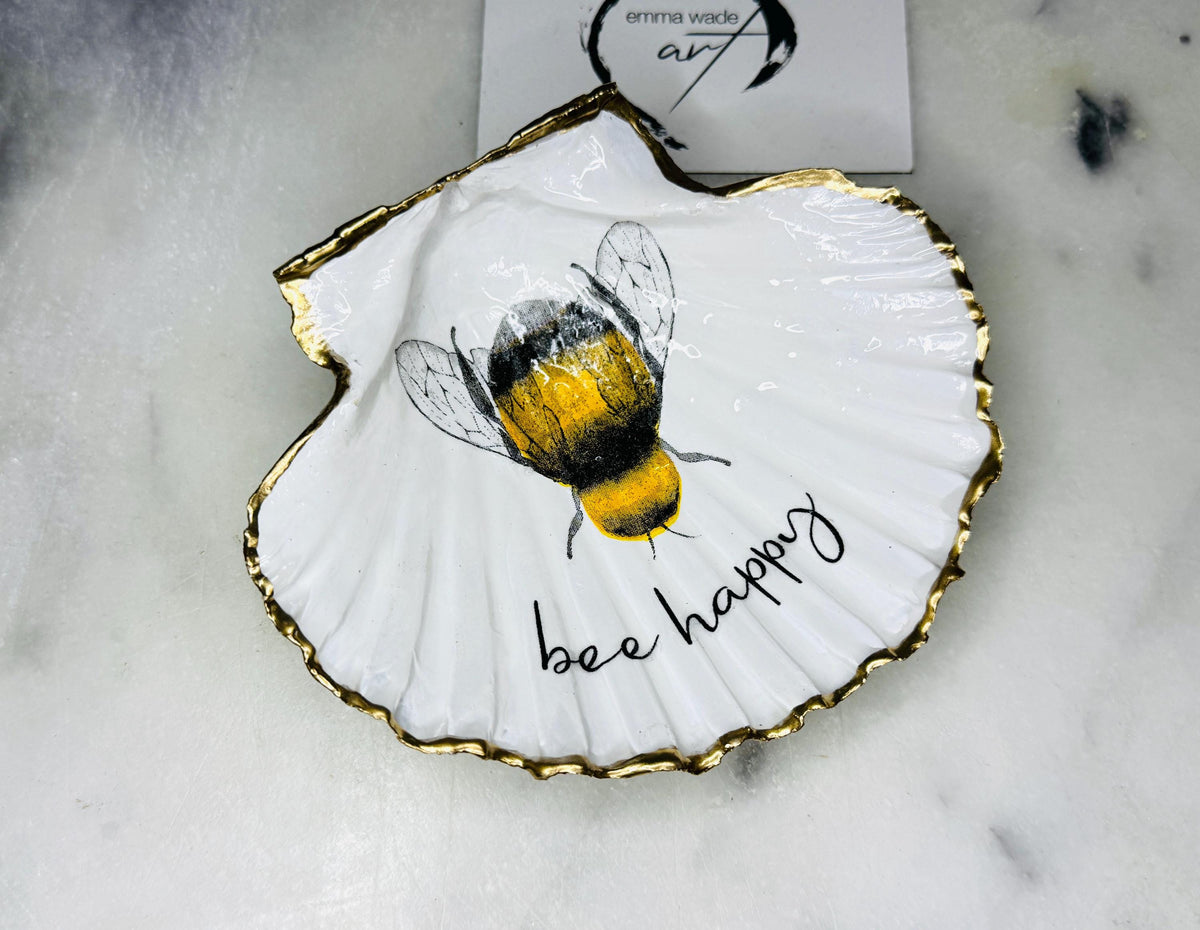 Handmade scallop shell trinket dish featuring a bumblebee illustration with gold leaf edging and glossy resin finish. Coastal jewellery dish in natural white shell with bee artwork, displayed as a ring holder and handmade bee lover gift