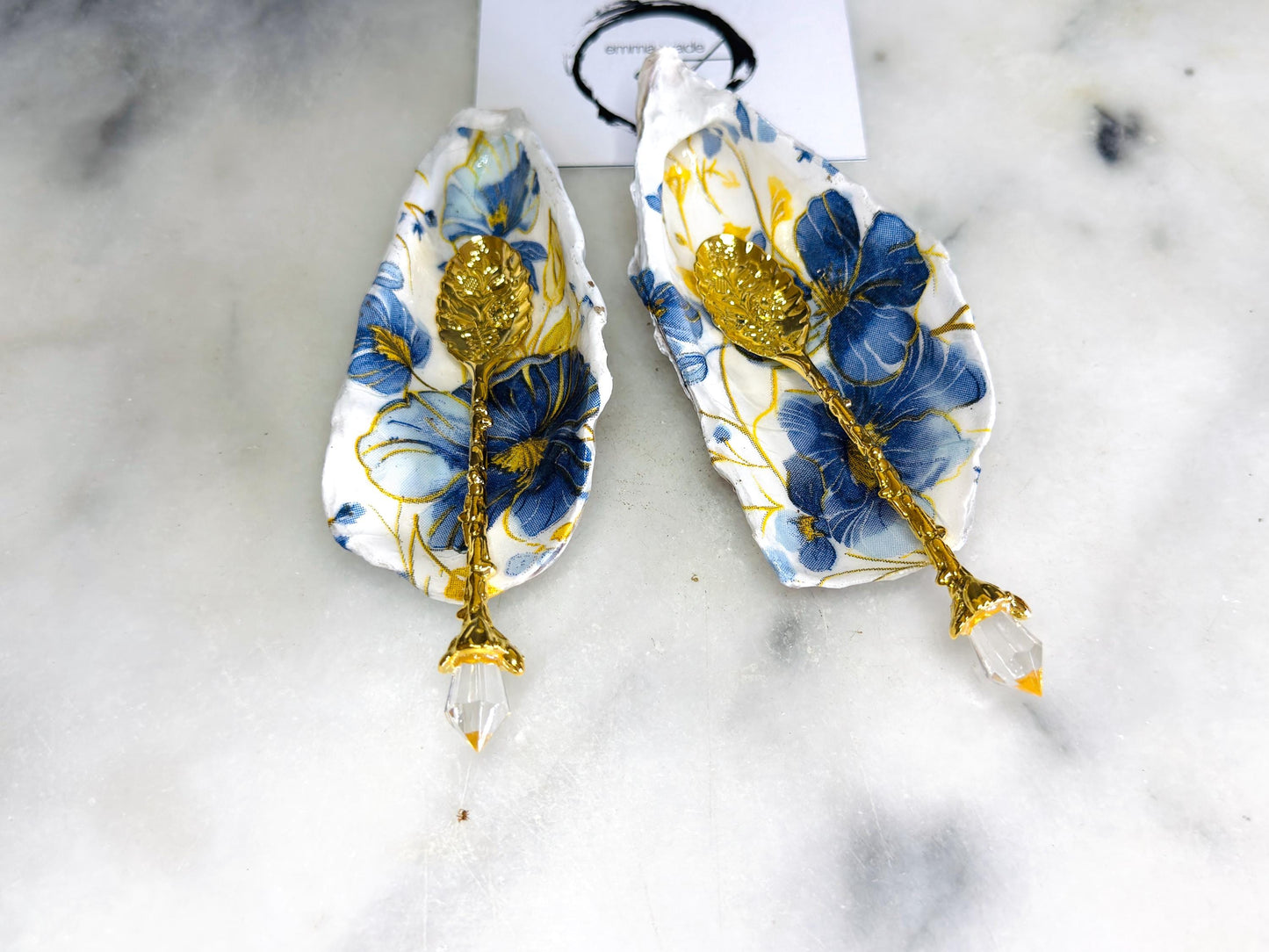 Handmade blue and yellow floral oyster shell salt and pepper dishes shown as a matching pair, crafted from natural Dorset shells and styled with small gold spoons. Coastal-inspired pinch pots featuring a decorative floral design with natural shell backs, perfect for table decor, weddings, or a unique hostess gift. Each shell is one of a kind, finished by hand, and ideal for adding colour and elegance to a dining table.