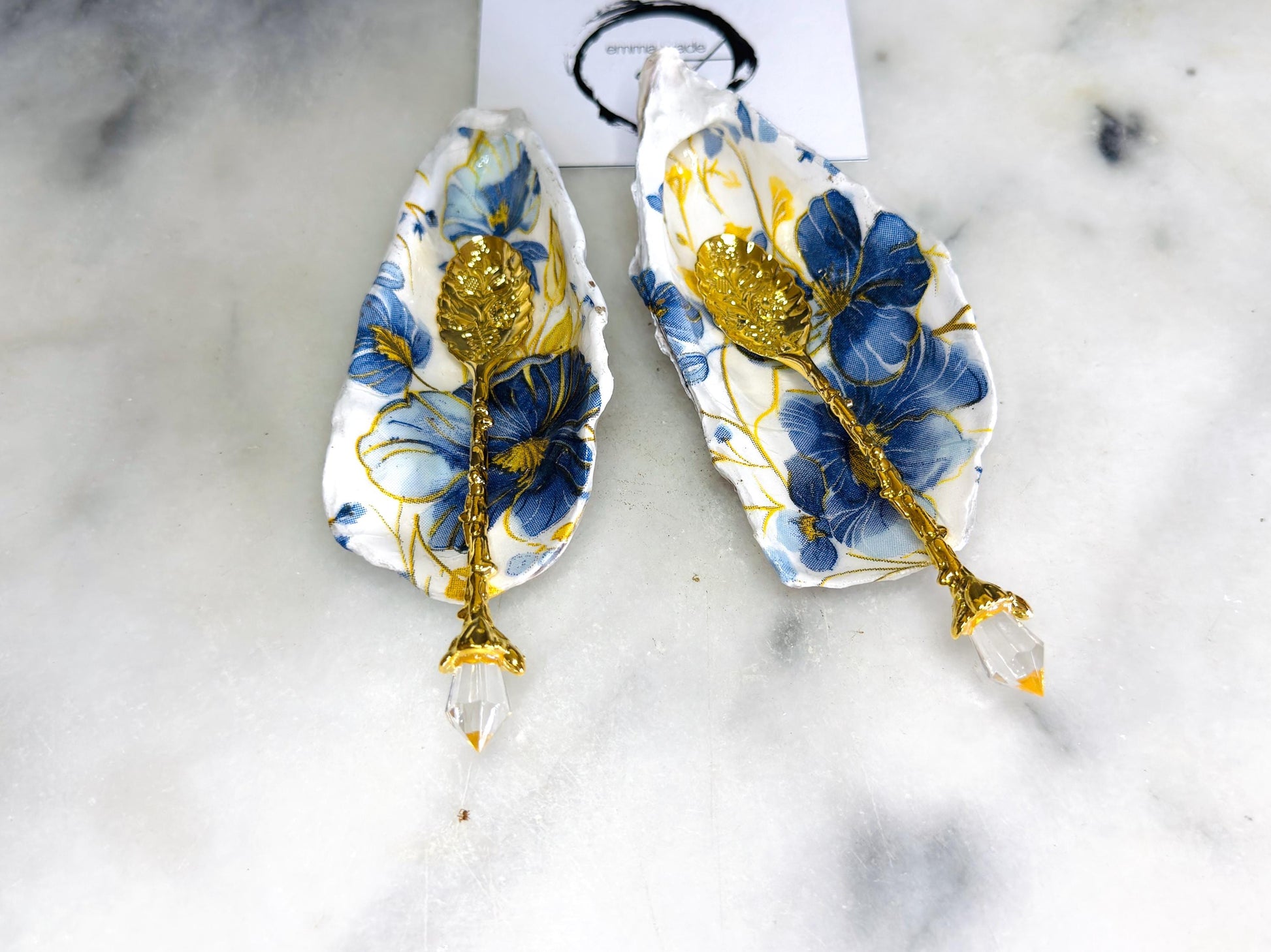 Handmade blue and yellow floral oyster shell salt and pepper dishes shown as a matching pair, crafted from natural Dorset shells and styled with small gold spoons. Coastal-inspired pinch pots featuring a decorative floral design with natural shell backs, perfect for table decor, weddings, or a unique hostess gift. Each shell is one of a kind, finished by hand, and ideal for adding colour and elegance to a dining table.