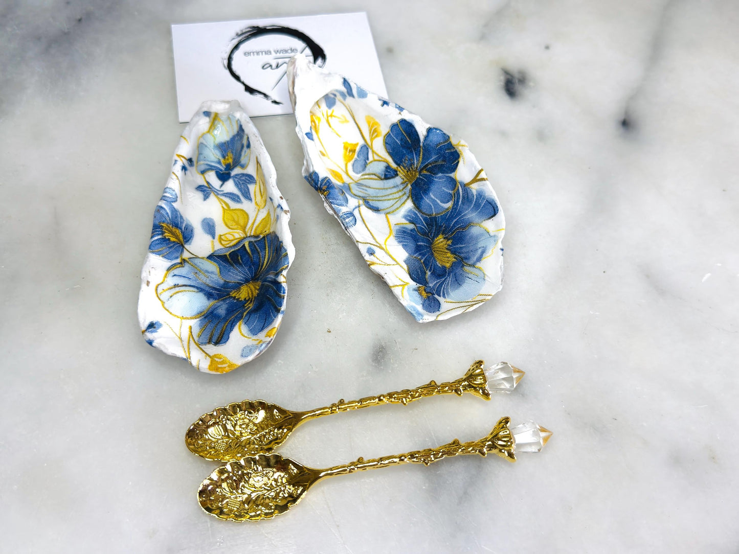 Handmade blue and yellow floral oyster shell salt and pepper dishes shown as a matching pair, crafted from natural Dorset shells and styled with small gold spoons. Coastal-inspired pinch pots featuring a decorative floral design with natural shell backs, perfect for table decor, weddings, or a unique hostess gift. Each shell is one of a kind, finished by hand, and ideal for adding colour and elegance to a dining table.