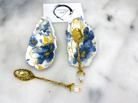 Handmade blue and yellow floral oyster shell salt and pepper dishes shown as a matching pair, crafted from natural Dorset shells and styled with small gold spoons. Coastal-inspired pinch pots featuring a decorative floral design with natural shell backs, perfect for table decor, weddings, or a unique hostess gift. Each shell is one of a kind, finished by hand, and ideal for adding colour and elegance to a dining table.
