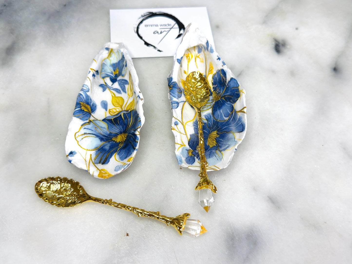 Handmade blue and yellow floral oyster shell salt and pepper dishes shown as a matching pair, crafted from natural Dorset shells and styled with small gold spoons. Coastal-inspired pinch pots featuring a decorative floral design with natural shell backs, perfect for table decor, weddings, or a unique hostess gift. Each shell is one of a kind, finished by hand, and ideal for adding colour and elegance to a dining table.