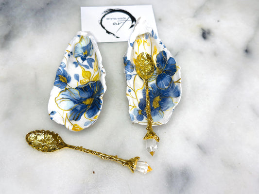 Handmade blue and yellow floral oyster shell salt and pepper dishes shown as a matching pair, crafted from natural Dorset shells and styled with small gold spoons. Coastal-inspired pinch pots featuring a decorative floral design with natural shell backs, perfect for table decor, weddings, or a unique hostess gift. Each shell is one of a kind, finished by hand, and ideal for adding colour and elegance to a dining table.
