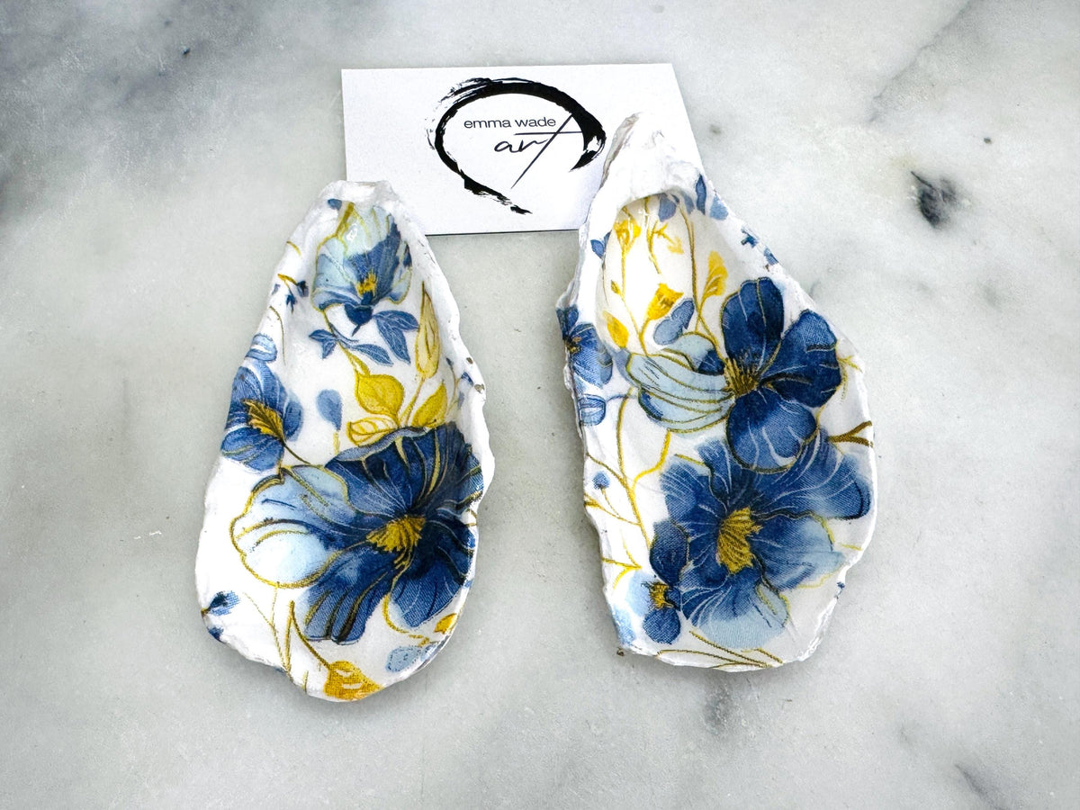 Handmade blue and yellow floral oyster shell salt and pepper dishes shown as a matching pair, crafted from natural Dorset shells and styled with small gold spoons. Coastal-inspired pinch pots featuring a decorative floral design with natural shell backs, perfect for table decor, weddings, or a unique hostess gift. Each shell is one of a kind, finished by hand, and ideal for adding colour and elegance to a dining table.