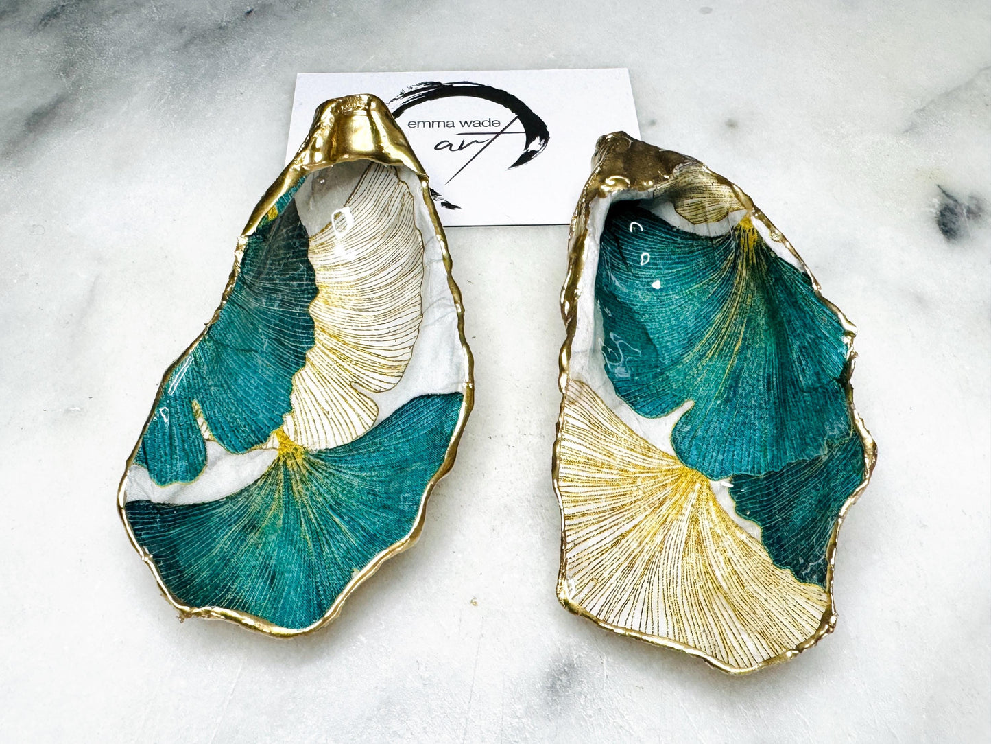 A pair of green and gold Japanese Ginkgo oyster shell salt and pepper pinch pots with floral botanical leaf design, hand gilded with liquid gold leaf and sealed in glossy resin. Coastal Dorset oyster shell dishes styled with matching gold crystal spoons on a marble background. Handmade luxury coastal dining décor and wedding gift.
