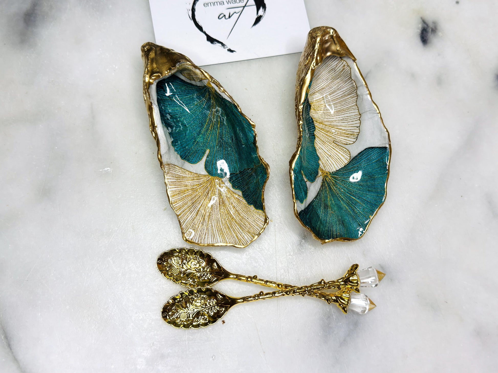 A pair of green and gold Japanese Ginkgo oyster shell salt and pepper pinch pots with floral botanical leaf design, hand gilded with liquid gold leaf and sealed in glossy resin. Coastal Dorset oyster shell dishes styled with matching gold crystal spoons on a marble background. Handmade luxury coastal dining décor and wedding gift.