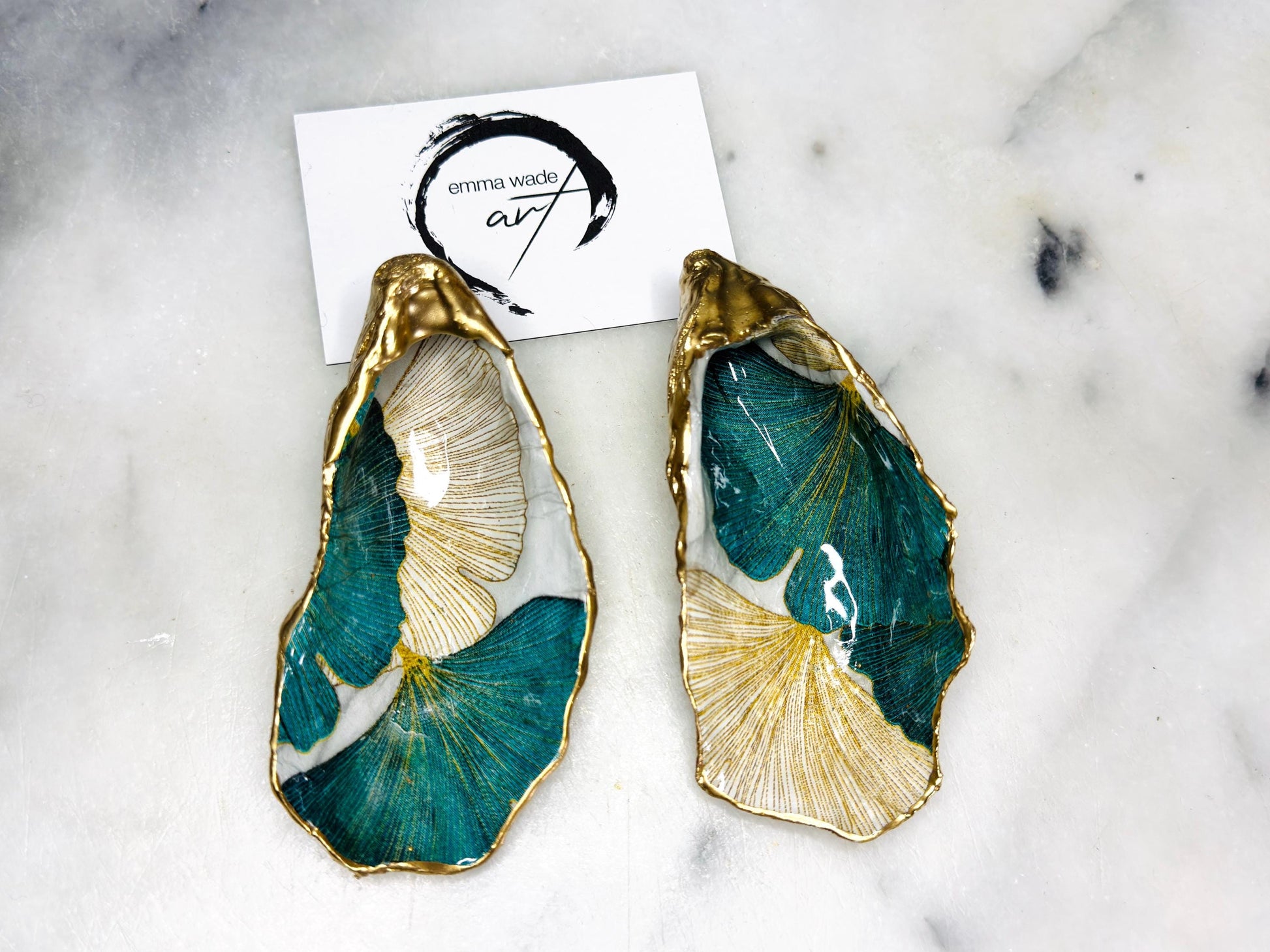 A pair of green and gold Japanese Ginkgo oyster shell salt and pepper pinch pots with floral botanical leaf design, hand gilded with liquid gold leaf and sealed in glossy resin. Coastal Dorset oyster shell dishes styled with matching gold crystal spoons on a marble background. Handmade luxury coastal dining décor and wedding gift.
