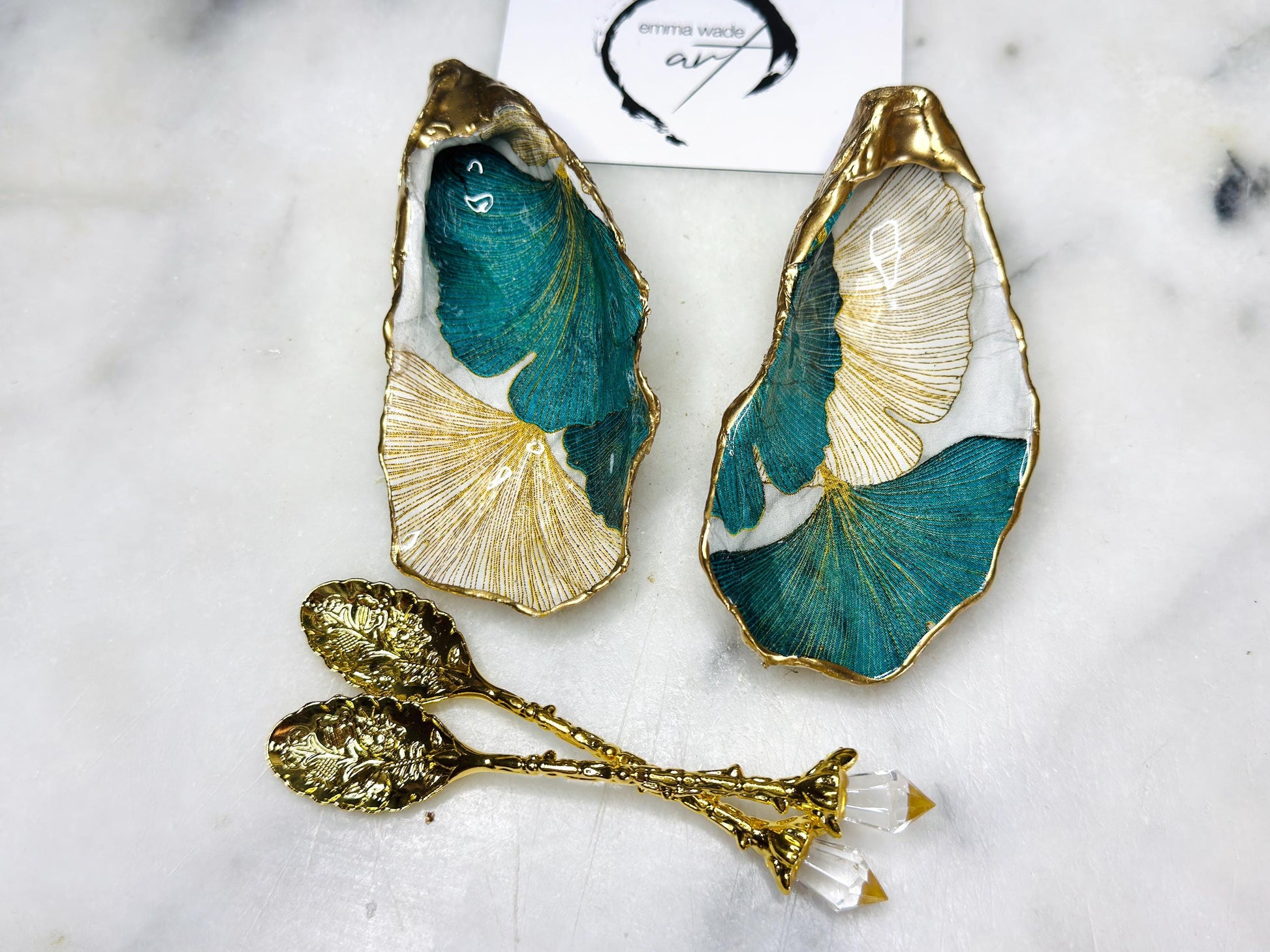 A pair of green and gold Japanese Ginkgo oyster shell salt and pepper pinch pots with floral botanical leaf design, hand gilded with liquid gold leaf and sealed in glossy resin. Coastal Dorset oyster shell dishes styled with matching gold crystal spoons on a marble background. Handmade luxury coastal dining décor and wedding gift.