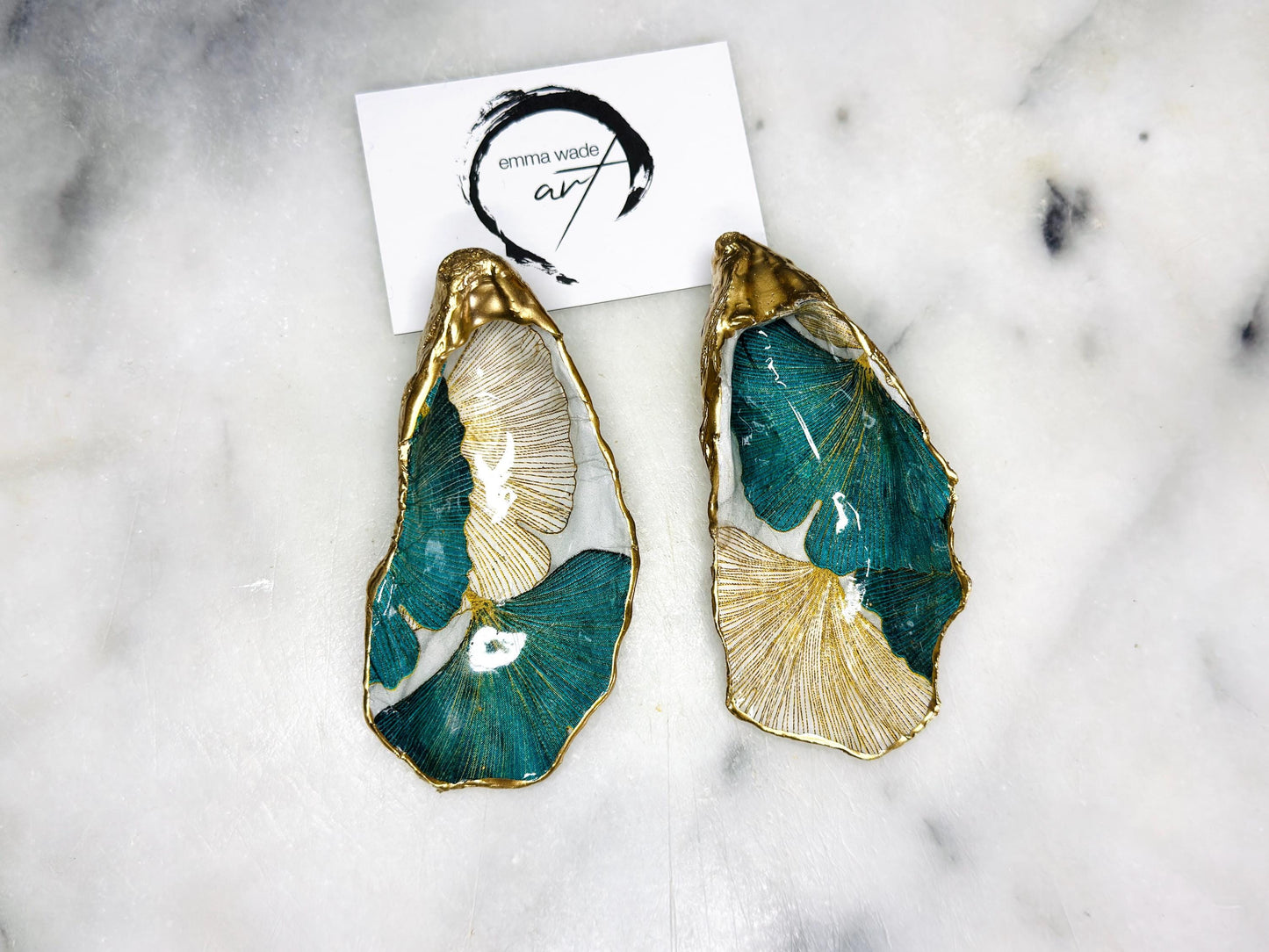 Handmade Dorset oyster shell ring dish featuring Japanese Ginkgo floral leaf artwork in emerald green, gold, and pearl tones with hand-applied liquid gold leaf edging. Luxury coastal jewellery tray sealed in glossy crystal-clear epoxy resin, styled with gold rings.