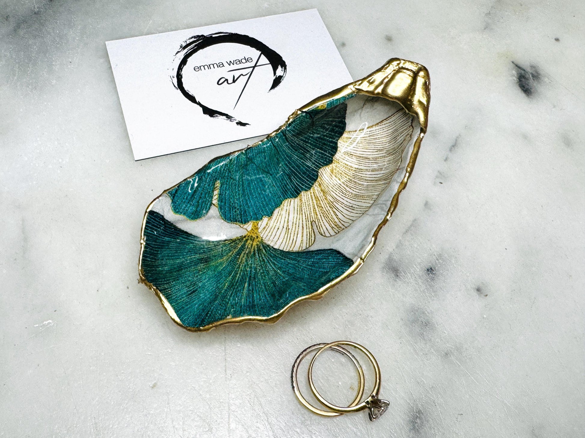 Handmade Dorset oyster shell ring dish featuring Japanese Ginkgo floral leaf artwork in emerald green, gold, and pearl tones with hand-applied liquid gold leaf edging. Luxury coastal jewellery tray sealed in glossy crystal-clear epoxy resin, styled with gold rings.