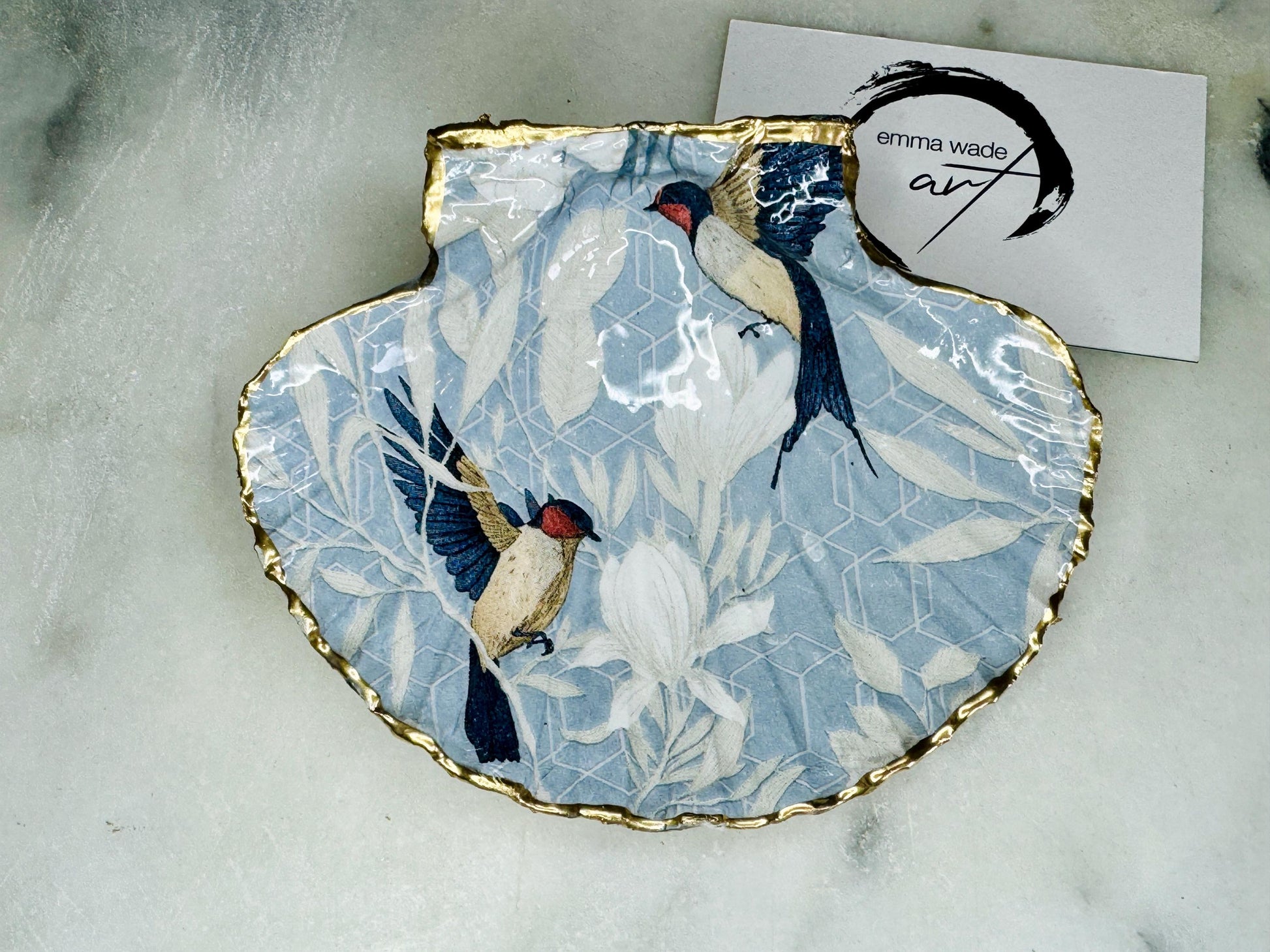 Handmade scallop shell trinket dish featuring a Blue Birds of Paradise decoupage design, finished with real gold leaf and sealed with glossy resin. Crafted from a natural Dorset scallop shell and shown as a jewellery and ring holder on a light background. Coastal-inspired home decor, perfect for holding rings, earrings and small keepsakes, with each shell one of a kind and finished by hand.