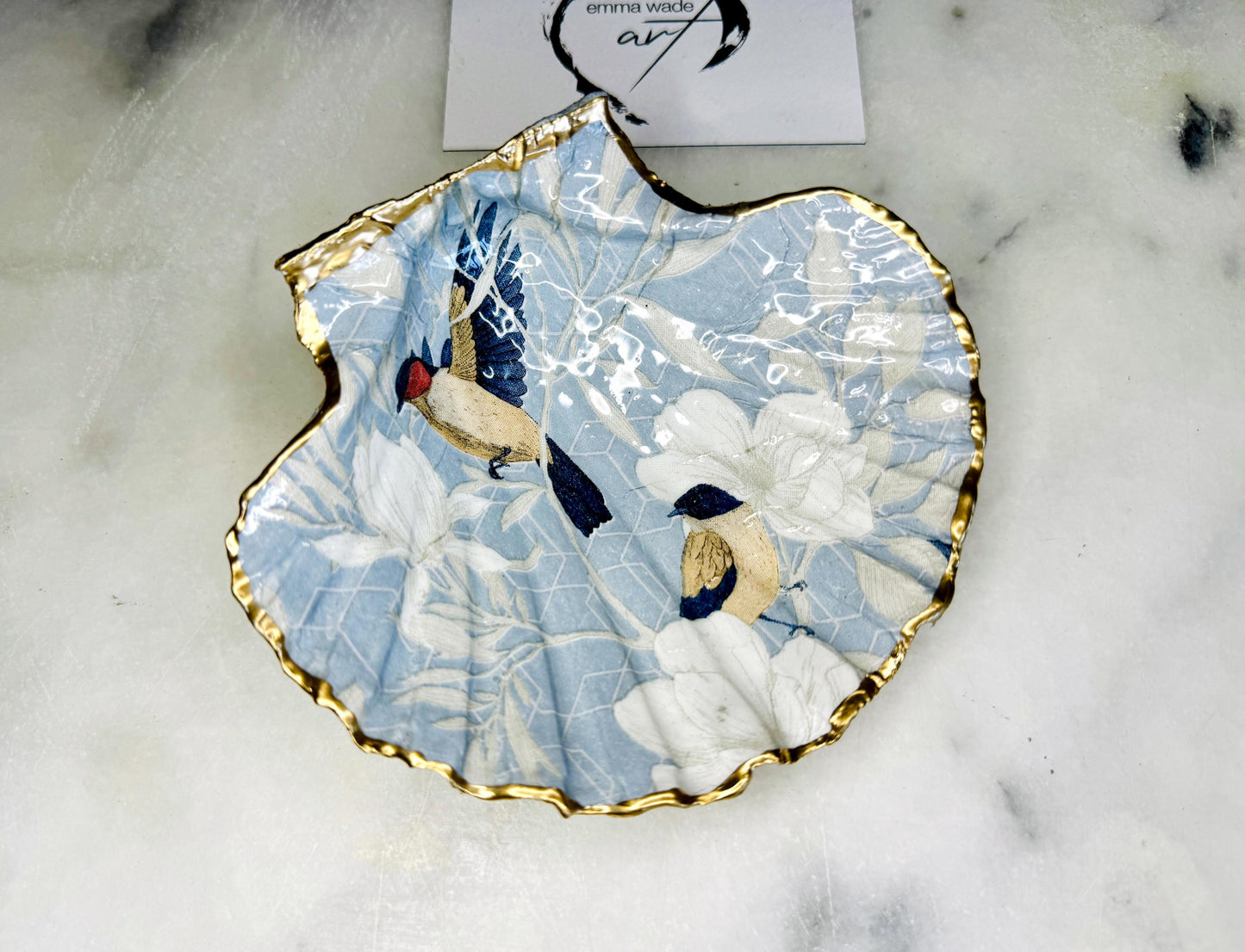 Handmade scallop shell trinket dish featuring a Blue Birds of Paradise decoupage design, finished with real gold leaf and sealed with glossy resin. Crafted from a natural Dorset scallop shell and shown as a jewellery and ring holder on a light background. Coastal-inspired home decor, perfect for holding rings, earrings and small keepsakes, with each shell one of a kind and finished by hand.