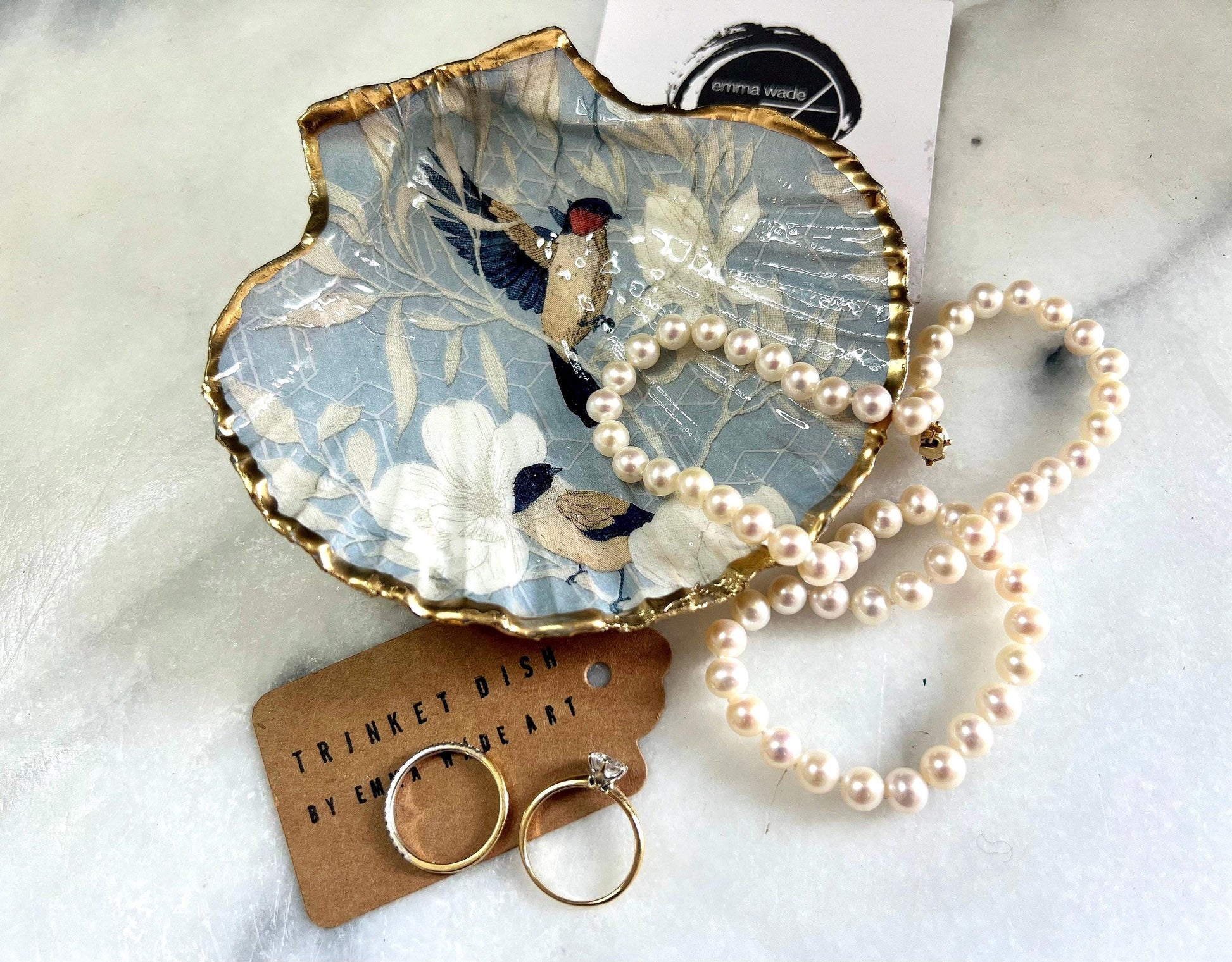 Handmade scallop shell trinket dish featuring a Blue Birds of Paradise decoupage design, finished with real gold leaf and sealed with glossy resin. Crafted from a natural Dorset scallop shell and shown as a jewellery and ring holder on a light background. Coastal-inspired home decor, perfect for holding rings, earrings and small keepsakes, with each shell one of a kind and finished by hand.