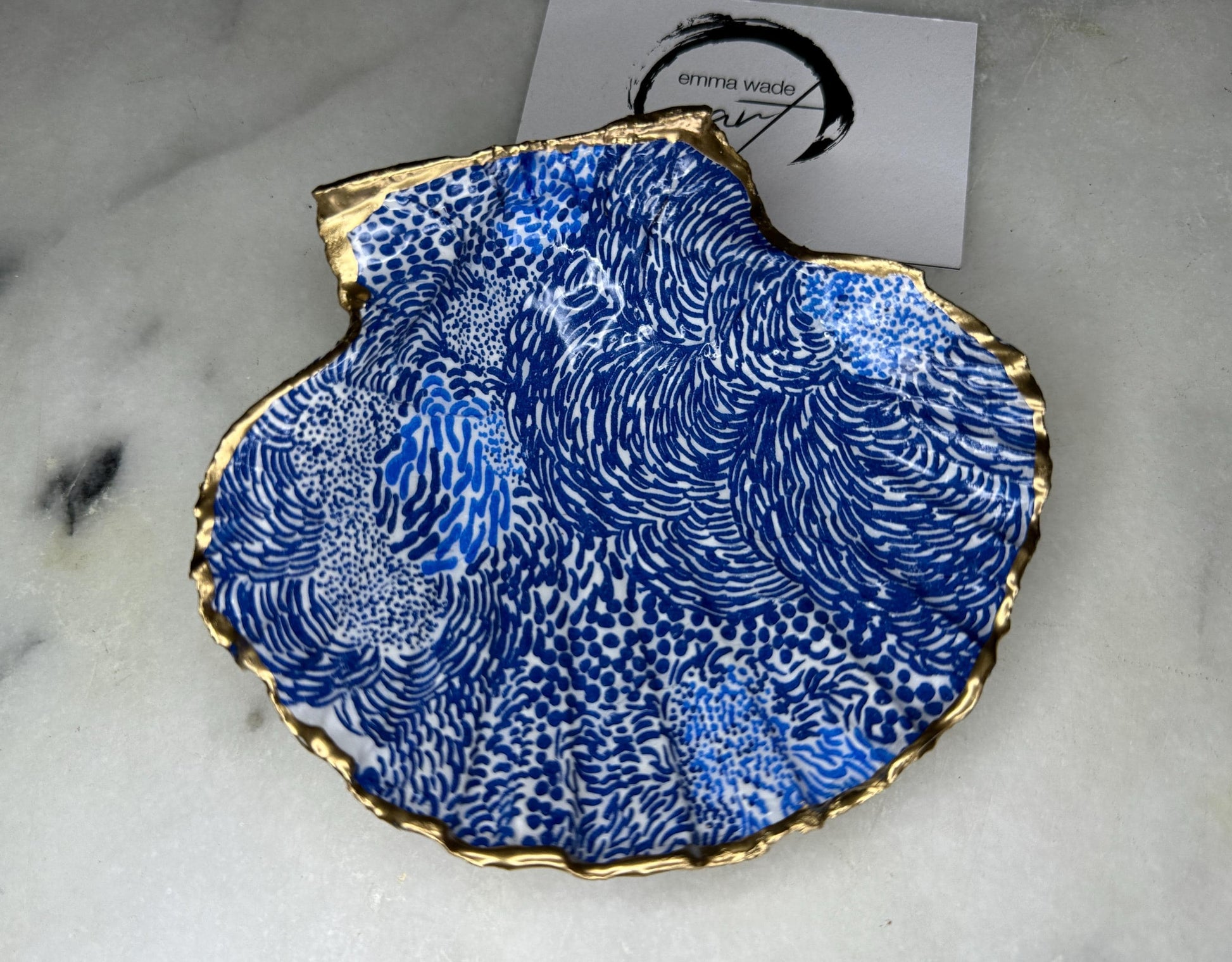 Marimekko inspired blue floral scallop shell trinket dish with hand-applied gold leaf edging and glossy resin finish. Natural Dorset shell jewellery holder featuring bold Scandinavian style botanical pattern. Decorative coastal ring dish styled on a marble surface, ideal as a bedside trinket bowl, host gift or luxury handmade home accent.
