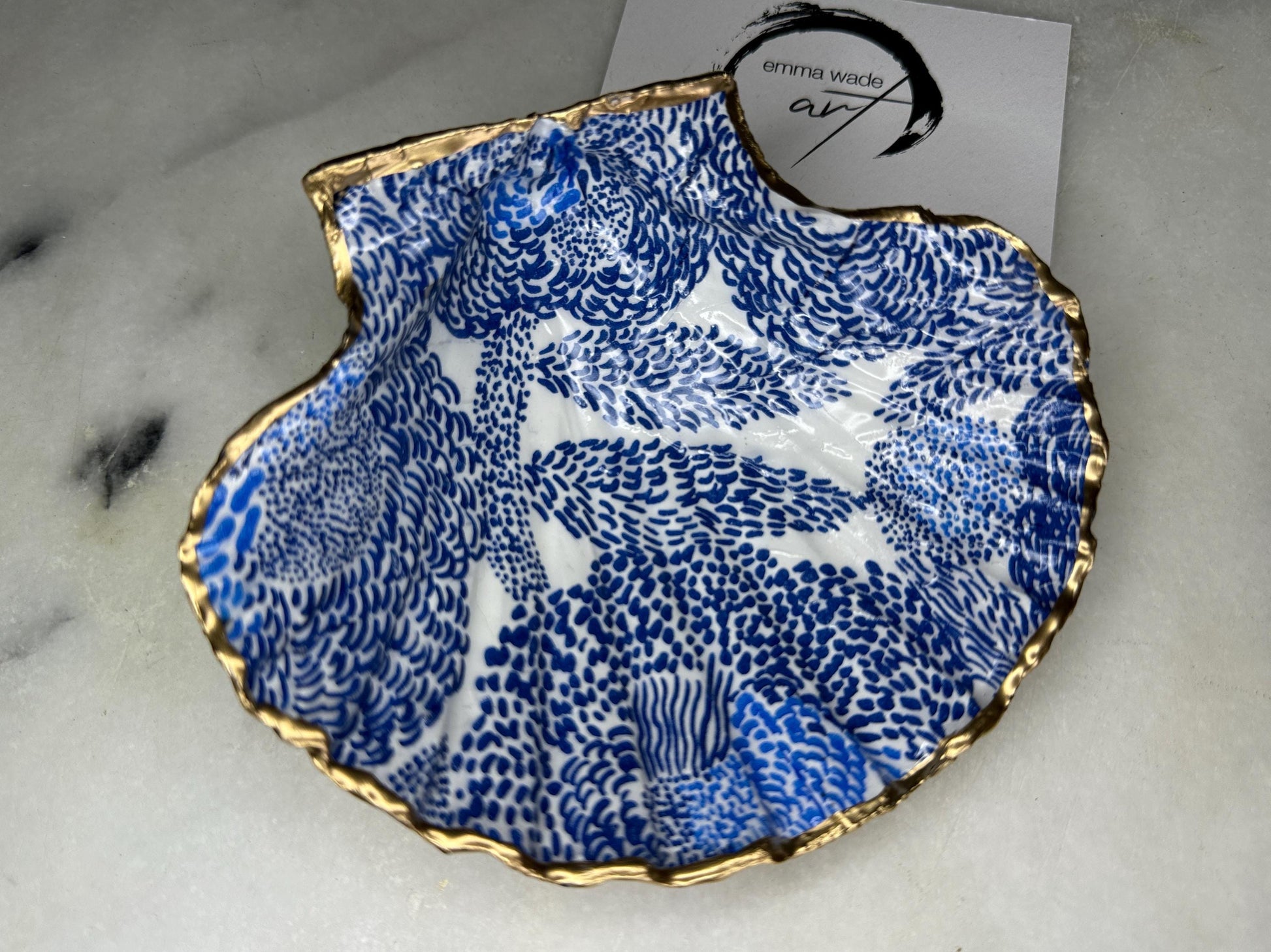 Marimekko inspired blue floral scallop shell trinket dish with hand-applied gold leaf edging and glossy resin finish. Natural Dorset shell jewellery holder featuring bold Scandinavian style botanical pattern. Decorative coastal ring dish styled on a marble surface, ideal as a bedside trinket bowl, host gift or luxury handmade home accent.
