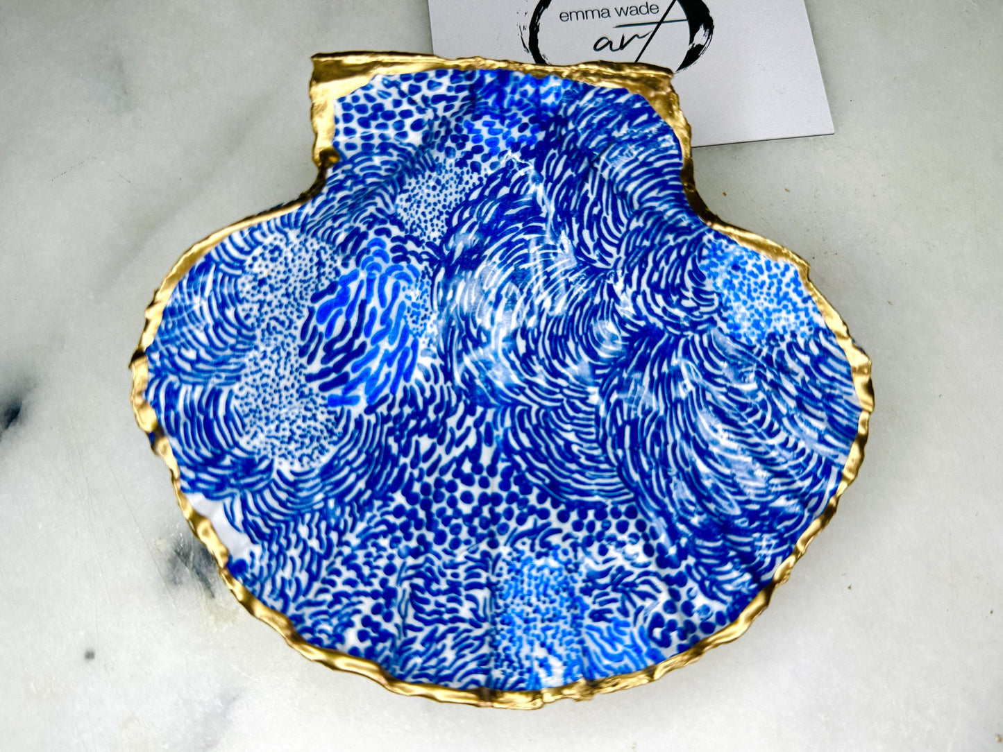 Marimekko inspired blue floral scallop shell trinket dish with hand-applied gold leaf edging and glossy resin finish. Natural Dorset shell jewellery holder featuring bold Scandinavian style botanical pattern. Decorative coastal ring dish styled on a marble surface, ideal as a bedside trinket bowl, host gift or luxury handmade home accent.