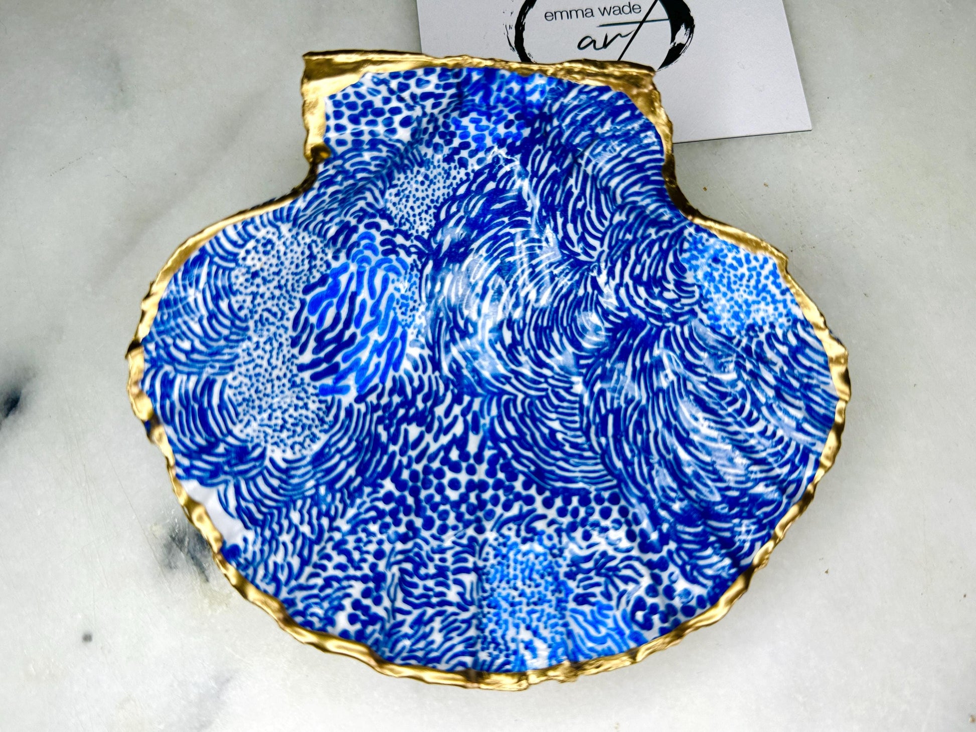 Marimekko inspired blue floral scallop shell trinket dish with hand-applied gold leaf edging and glossy resin finish. Natural Dorset shell jewellery holder featuring bold Scandinavian style botanical pattern. Decorative coastal ring dish styled on a marble surface, ideal as a bedside trinket bowl, host gift or luxury handmade home accent.
