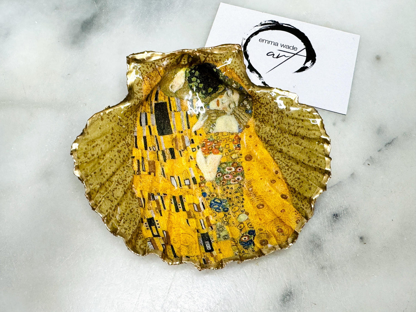 Handmade scallop shell trinket dish inspired by Gustav Klimt’s “The Kiss”, finished with gold leaf and sealed with glossy resin. Coastal Dorset shell jewellery dish perfect for rings, earrings and small treasures. Unique decorative shell bowl with golden artwork design, photographed on a light background showing detailed gilded surface and natural shell shape.