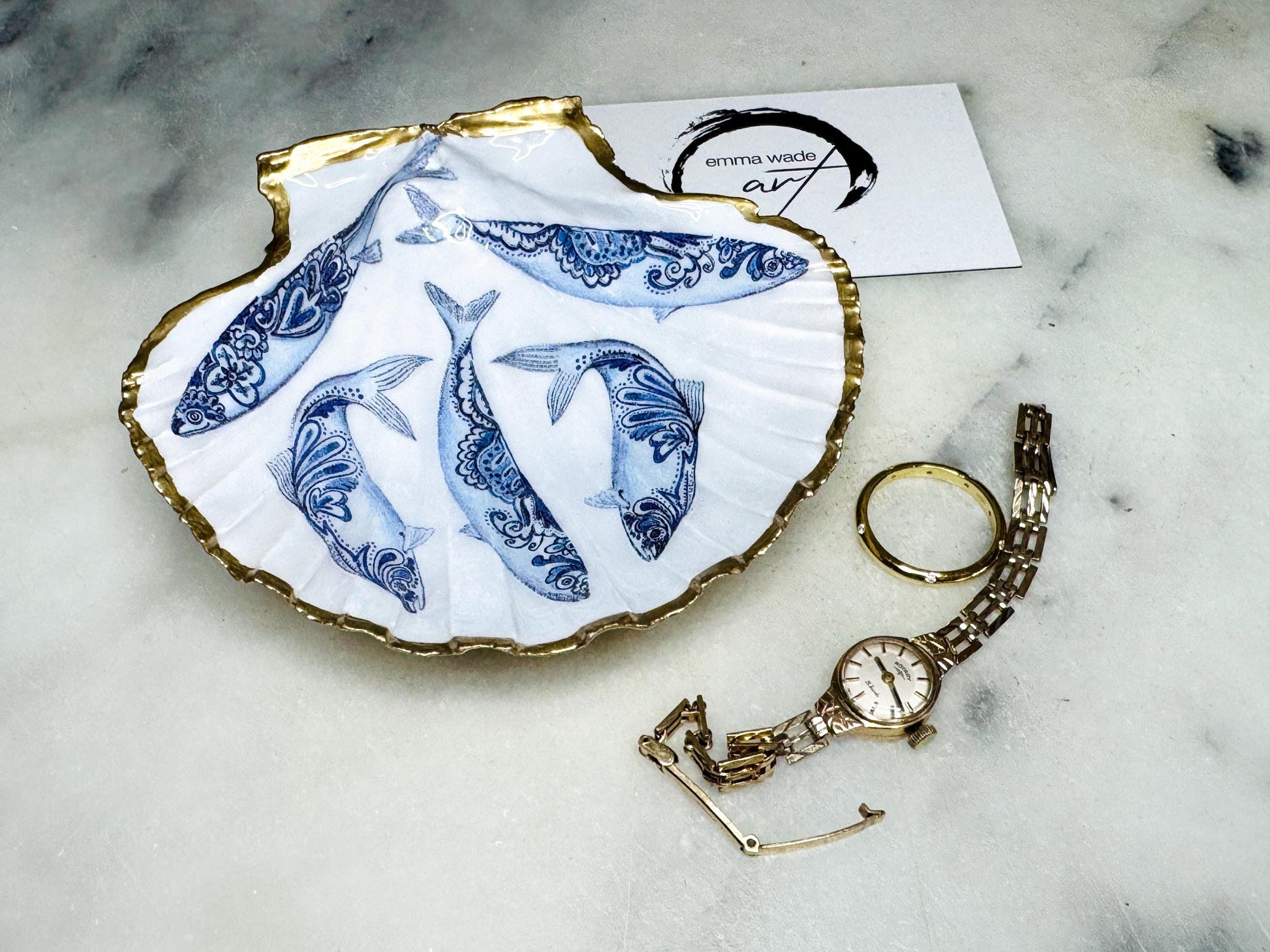 Handmade Dorset scallop shell trinket dishes featuring blue and white paisley fish illustrations with hand-painted gold leaf edging and glossy crystal clear resin. Coastal Scandinavian inspired jewellery dishes styled on a soft neutral background with elegant nautical fish artwork and luxurious gilded detailing.