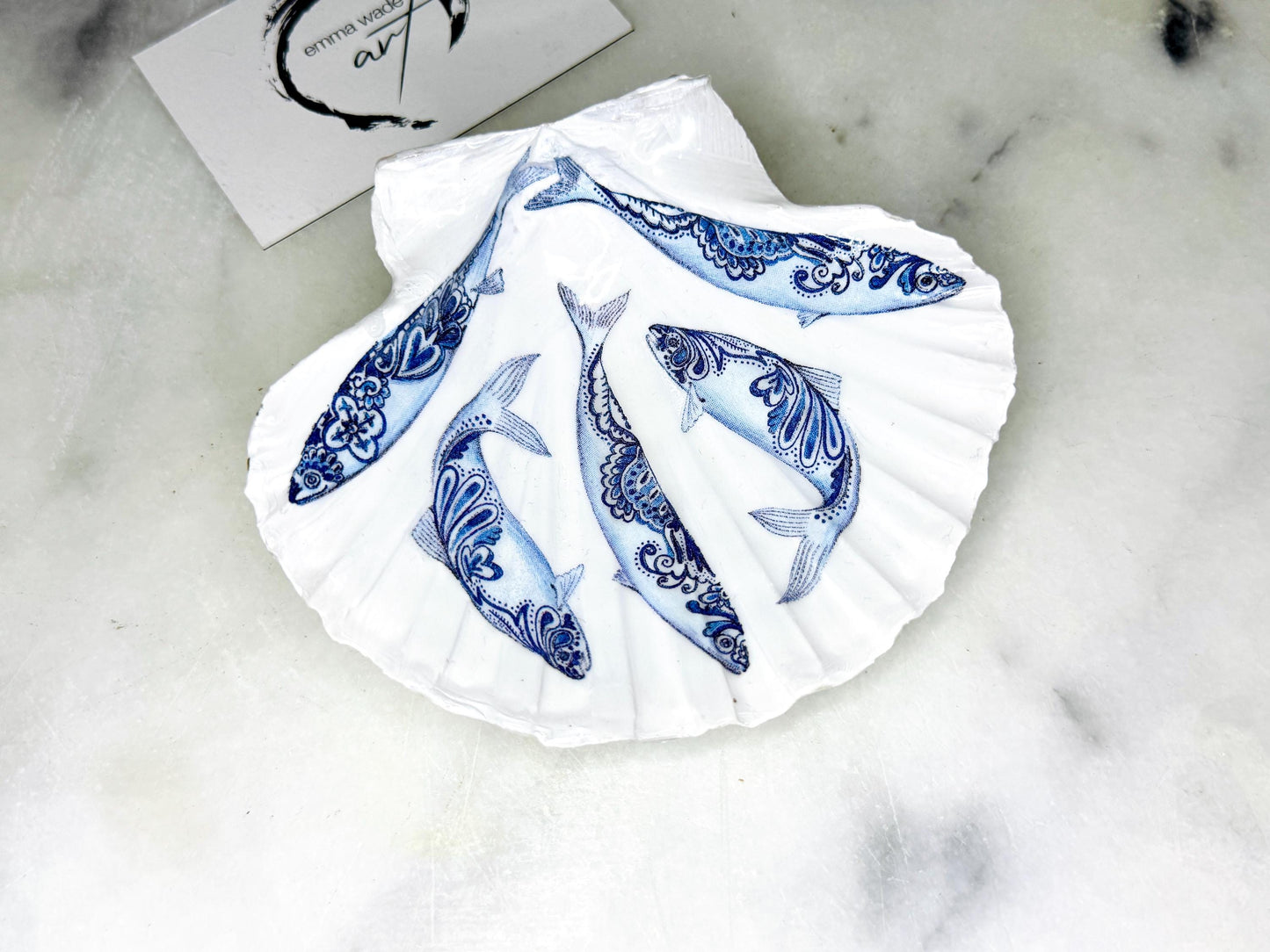 Handmade Dorset scallop shell trinket dishes featuring blue and white paisley fish illustrations with hand-painted gold leaf edging and glossy crystal clear resin. Coastal Scandinavian inspired jewellery dishes styled on a soft neutral background with elegant nautical fish artwork and luxurious gilded detailing.