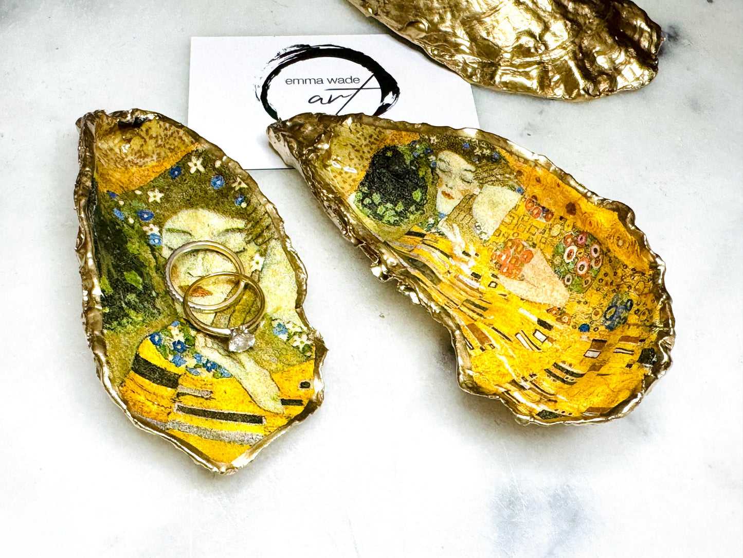 Handcrafted Dorset oyster shell trinket dish inspired by Gustav Klimt’s The Kiss, featuring romantic gold and green artwork detail. Natural oyster shell edged in hand applied liquid gold leaf and sealed with high gloss resin. Luxury art inspired jewellery holder displayed with gold rings on marble background. Unique gift for her or art lover keepsake.