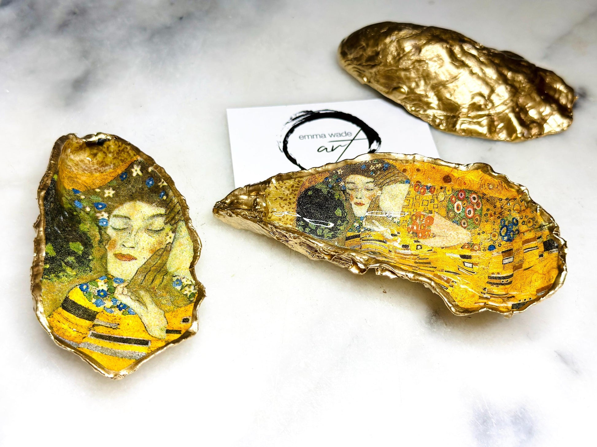 Handcrafted Dorset oyster shell trinket dish inspired by Gustav Klimt’s The Kiss, featuring romantic gold and green artwork detail. Natural oyster shell edged in hand applied liquid gold leaf and sealed with high gloss resin. Luxury art inspired jewellery holder displayed with gold rings on marble background. Unique gift for her or art lover keepsake.