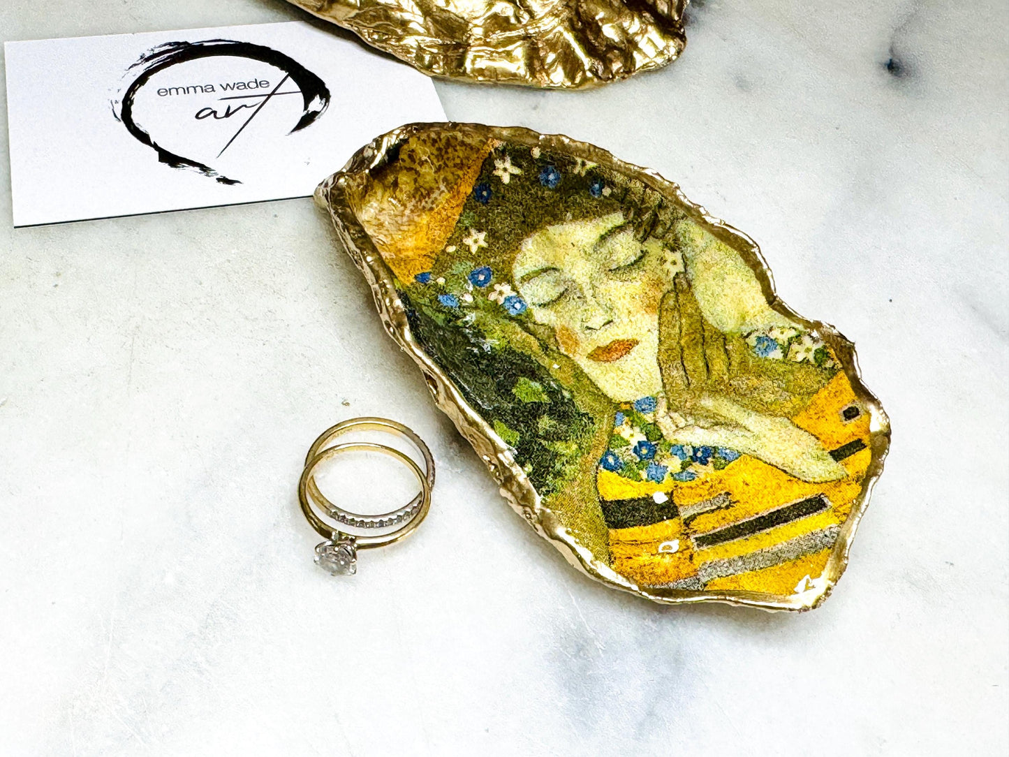 Handcrafted Dorset oyster shell trinket dish inspired by Gustav Klimt’s The Kiss, featuring romantic gold and green artwork detail. Natural oyster shell edged in hand applied liquid gold leaf and sealed with high gloss resin. Luxury art inspired jewellery holder displayed with gold rings on marble background. Unique gift for her or art lover keepsake.