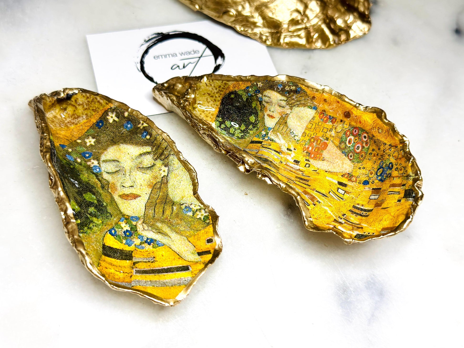 Handcrafted Dorset oyster shell trinket dish inspired by Gustav Klimt’s The Kiss, featuring romantic gold and green artwork detail. Natural oyster shell edged in hand applied liquid gold leaf and sealed with high gloss resin. Luxury art inspired jewellery holder displayed with gold rings on marble background. Unique gift for her or art lover keepsake.