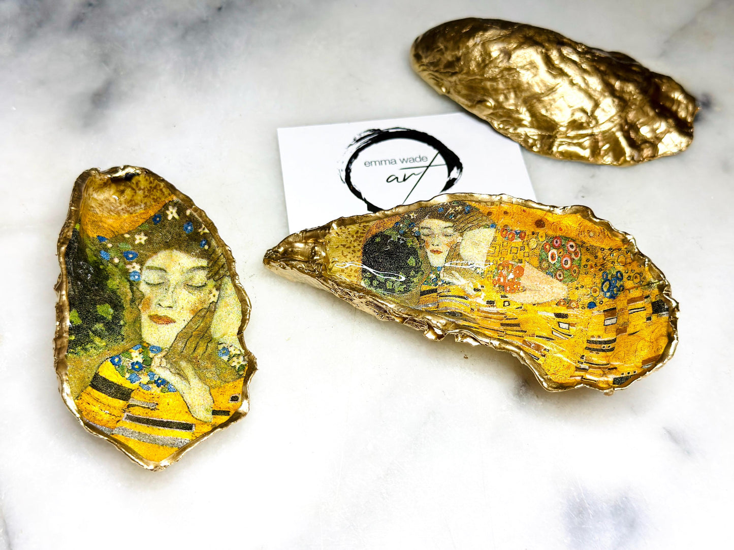 Handcrafted Dorset oyster shell trinket dish inspired by Gustav Klimt’s The Kiss, featuring romantic gold and green artwork detail. Natural oyster shell edged in hand applied liquid gold leaf and sealed with high gloss resin. Luxury art inspired jewellery holder displayed with gold rings on marble background. Unique gift for her or art lover keepsake.