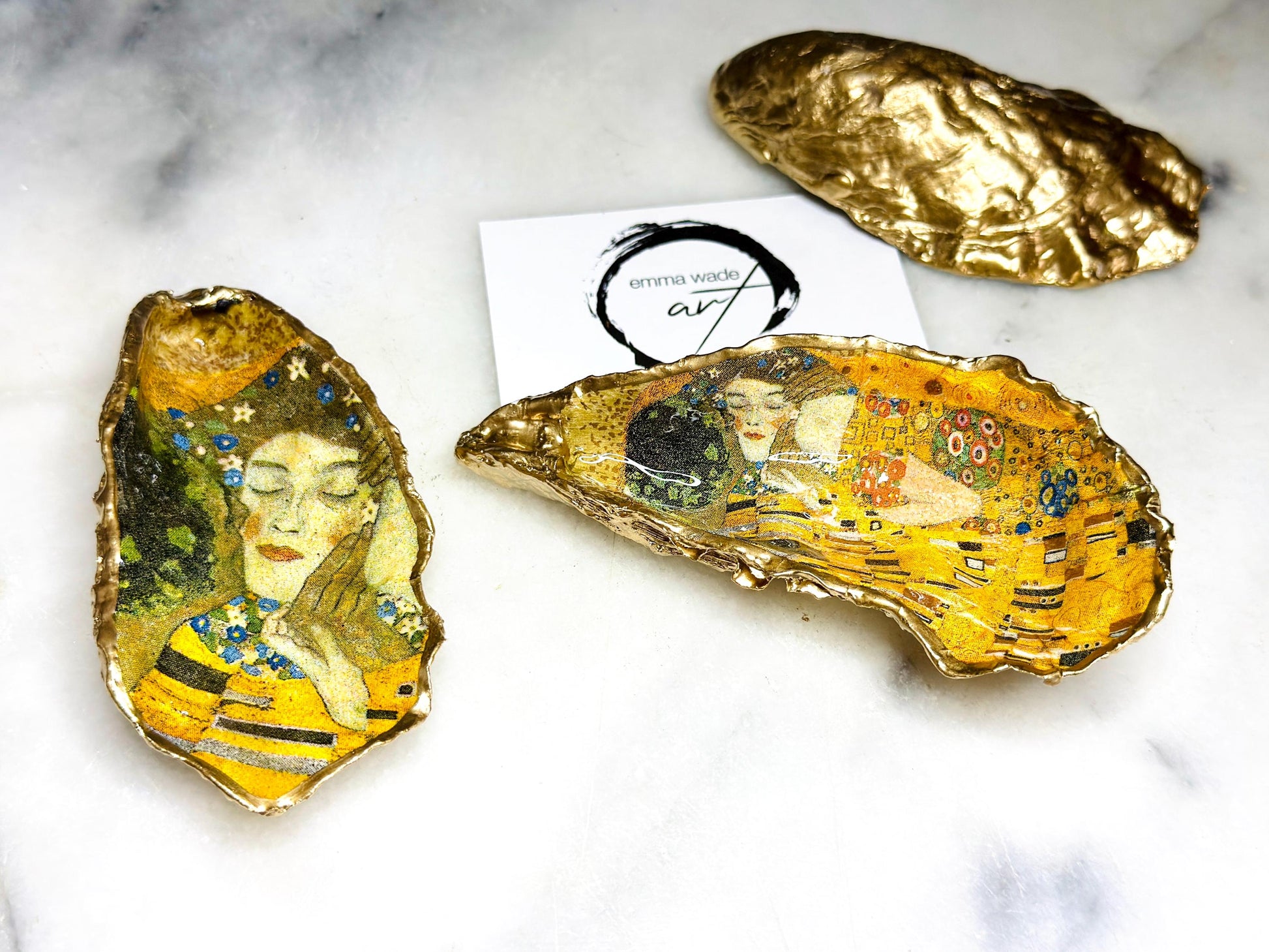 Handcrafted Dorset oyster shell trinket dish inspired by Gustav Klimt’s The Kiss, featuring romantic gold and green artwork detail. Natural oyster shell edged in hand applied liquid gold leaf and sealed with high gloss resin. Luxury art inspired jewellery holder displayed with gold rings on marble background. Unique gift for her or art lover keepsake.