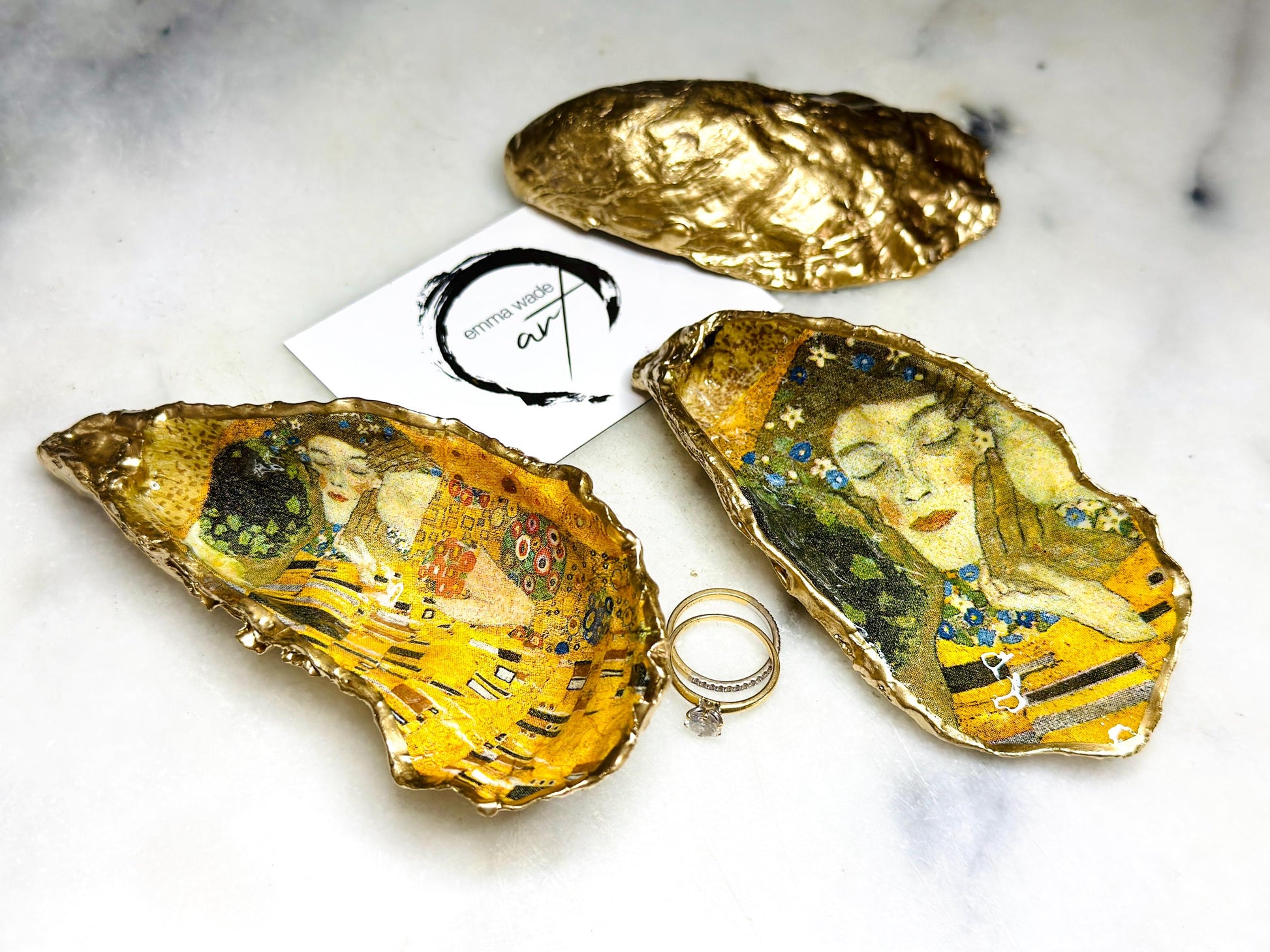 Handcrafted Dorset oyster shell trinket dish inspired by Gustav Klimt’s The Kiss, featuring romantic gold and green artwork detail. Natural oyster shell edged in hand applied liquid gold leaf and sealed with high gloss resin. Luxury art inspired jewellery holder displayed with gold rings on marble background. Unique gift for her or art lover keepsake.