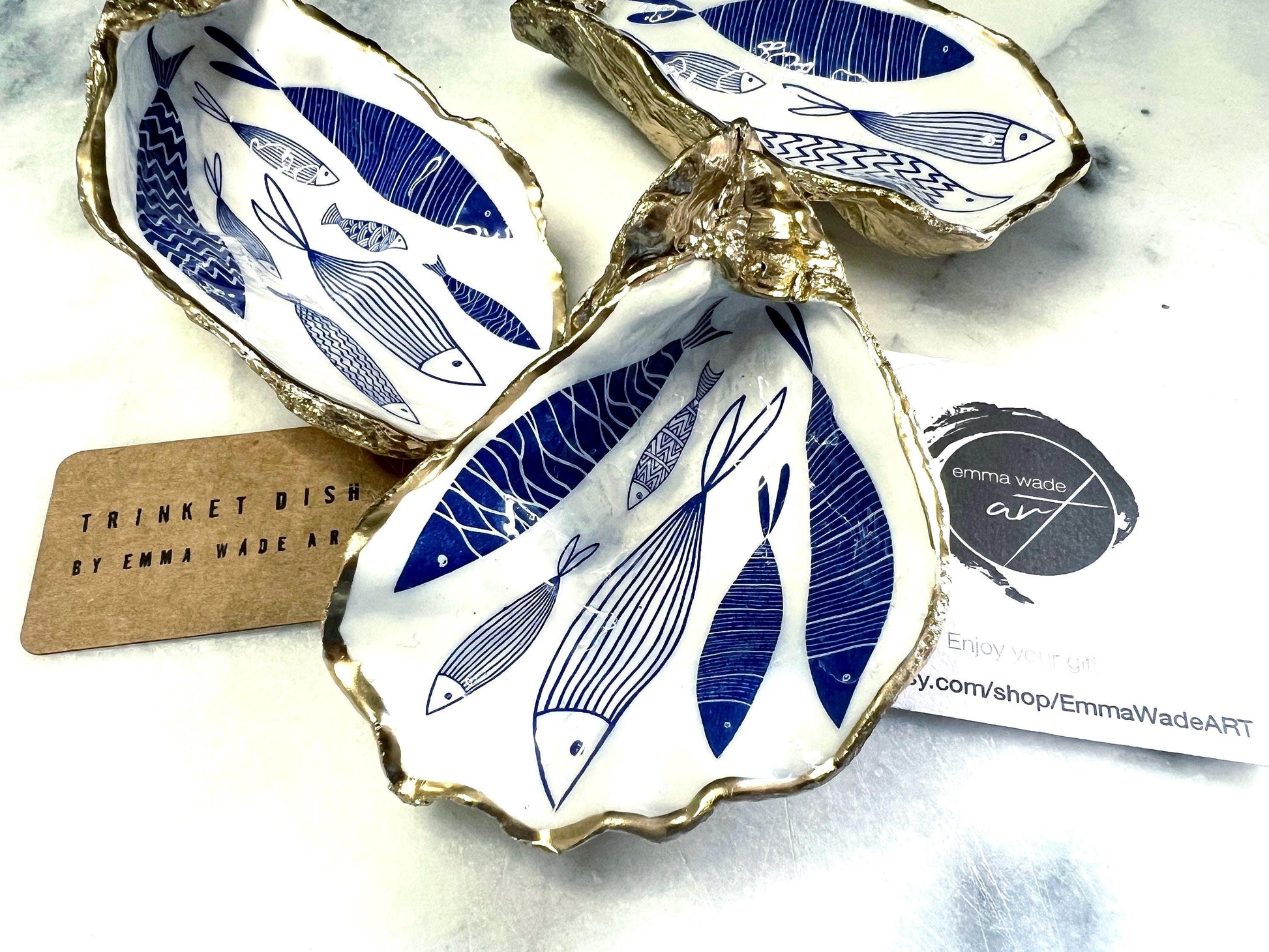 Hand-gilded Dorset oyster shell trinket dish featuring a blue fish motif, edged with liquid gold leaf and sealed in high gloss resin. Coastal ring holder and nautical home decor piece displayed with multiple shells. Sustainable oyster shell art handmade in Surrey, ideal as a jewellery dish, wedding favour, beach house accessory or ocean lover gift.