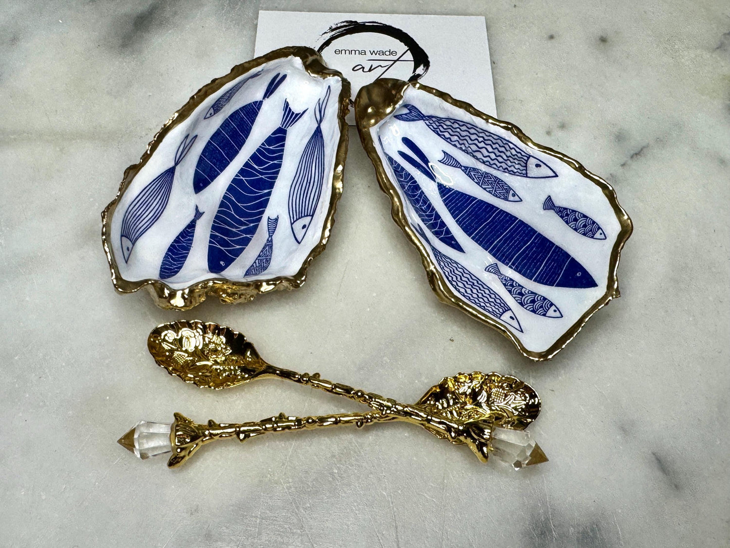 Pair of handmade Dorset oyster shell salt and pepper pinch pots decorated with elegant blue and white fish artwork, finished with hand applied liquid gold leaf edging and sealed in glossy food-safe resin. Coastal shell cruet dishes styled with optional gold crystal spoons on a marble background. Luxury nautical kitchen decor inspired by the sea, perfect for beach house dining, dinner parties, weddings, anniversaries, and coastal home gifts.
