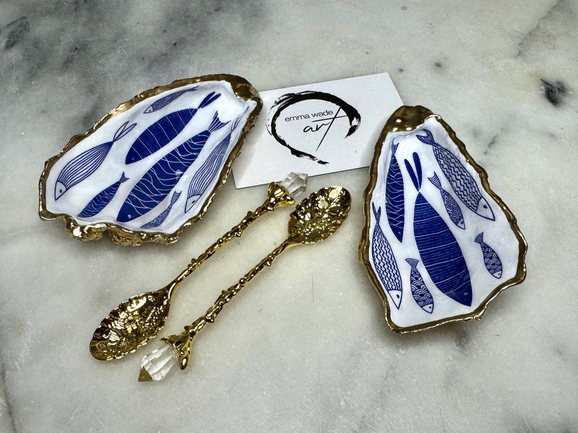 Pair of handmade Dorset oyster shell salt and pepper pinch pots decorated with elegant blue and white fish artwork, finished with hand applied liquid gold leaf edging and sealed in glossy food-safe resin. Coastal shell cruet dishes styled with optional gold crystal spoons on a marble background. Luxury nautical kitchen decor inspired by the sea, perfect for beach house dining, dinner parties, weddings, anniversaries, and coastal home gifts.